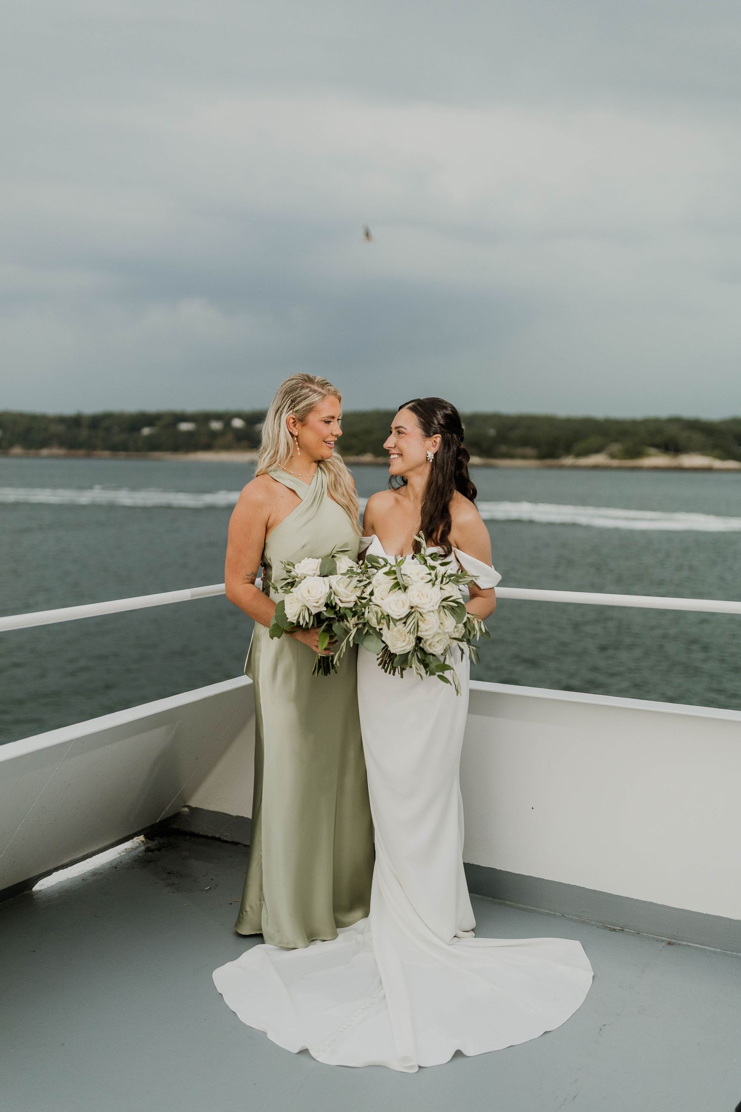 Bride in white gown and bridesmaid in sage green dress posing on boat deck.