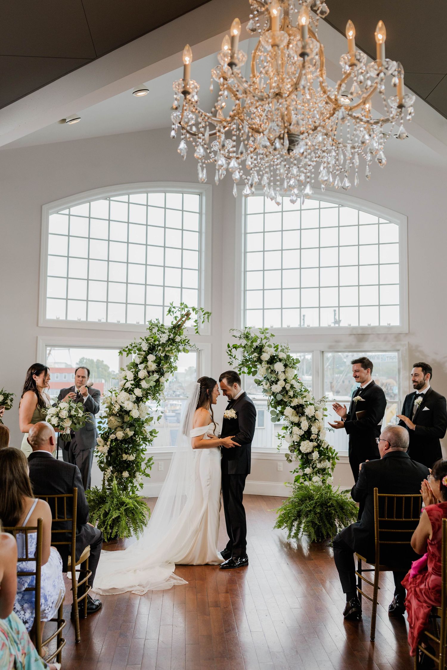 Bride and groom exchange vows under floral arch with white roses beneath crystal chandelier in elegant venue.