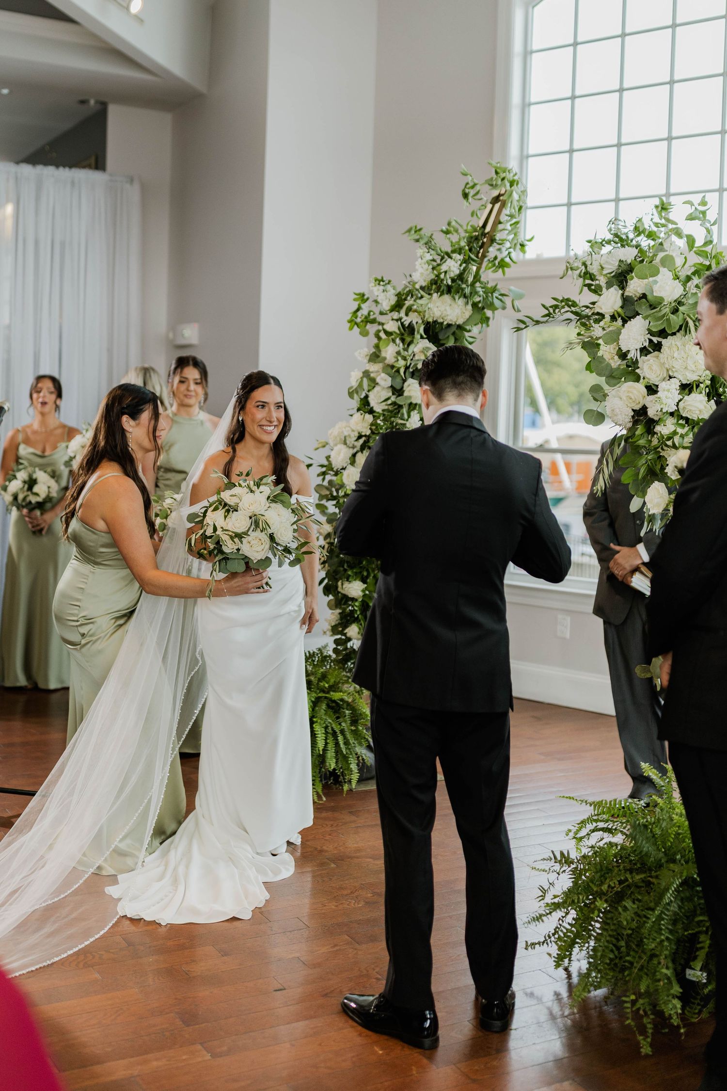 Bride in white gown smiling at groom during indoor wedding ceremony with white floral arch backdrop.