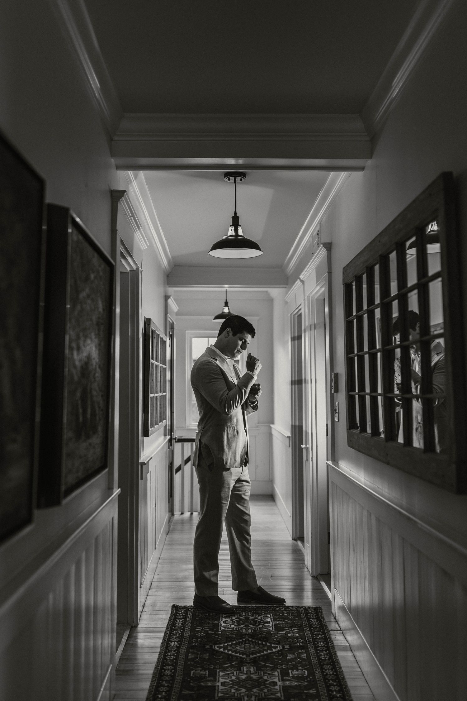 Black and white photo of groom adjusting tie in a hallway with framed artwork on walls.