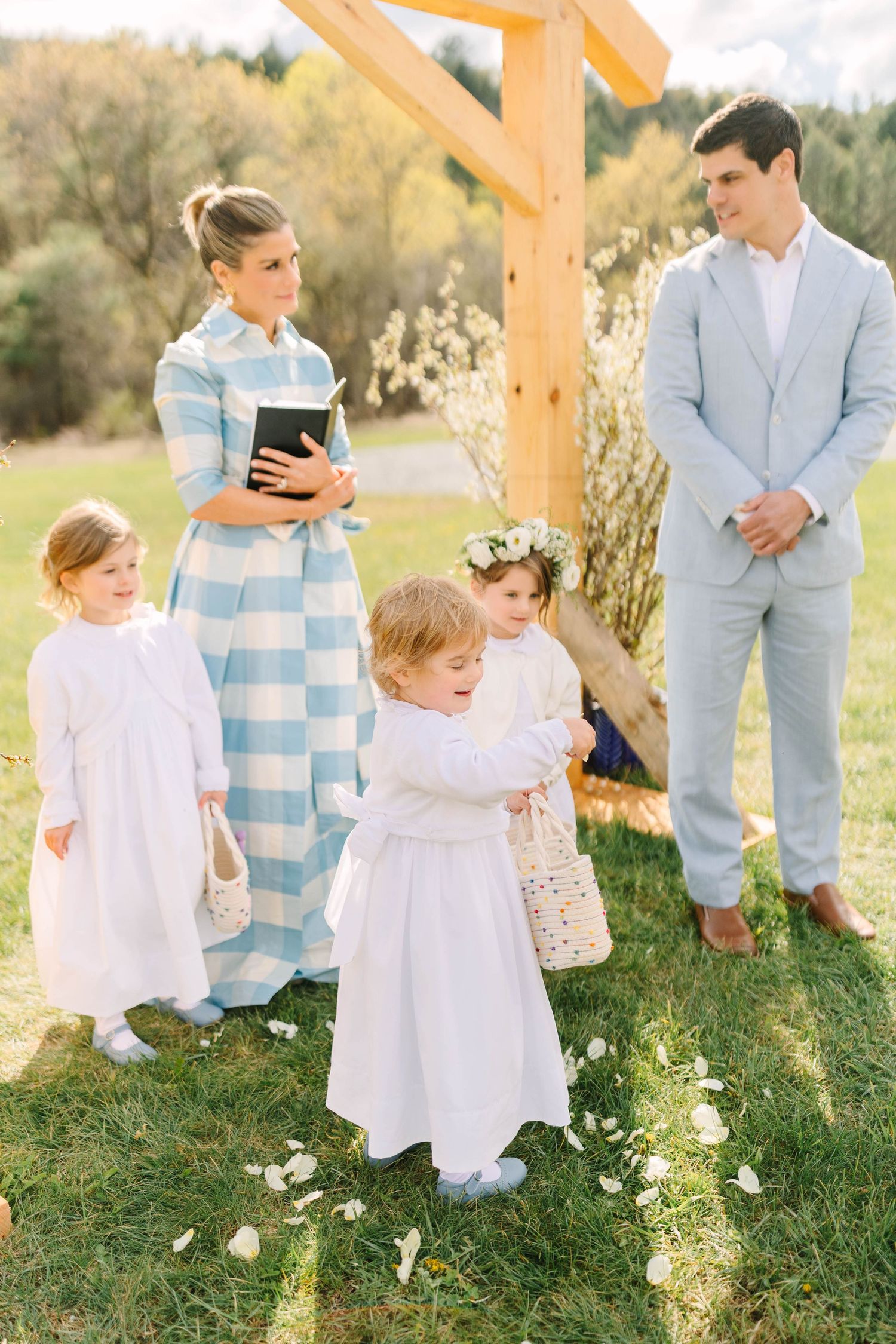 Flower girls in white dresses gather under wooden arch with officiant and groom at outdoor wedding.