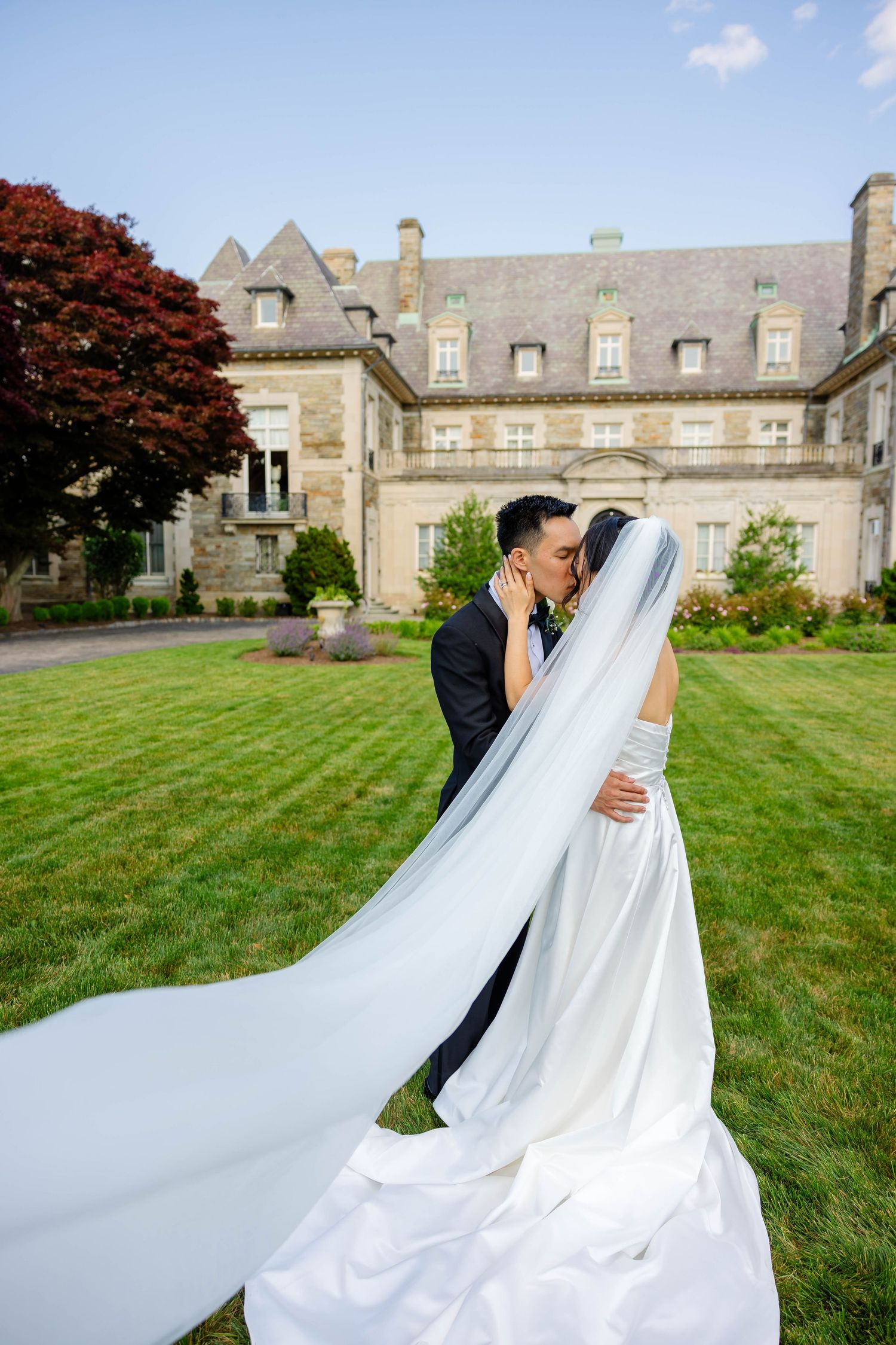 Bride and groom kiss on lush green lawn before grand stone manor house on wedding day.