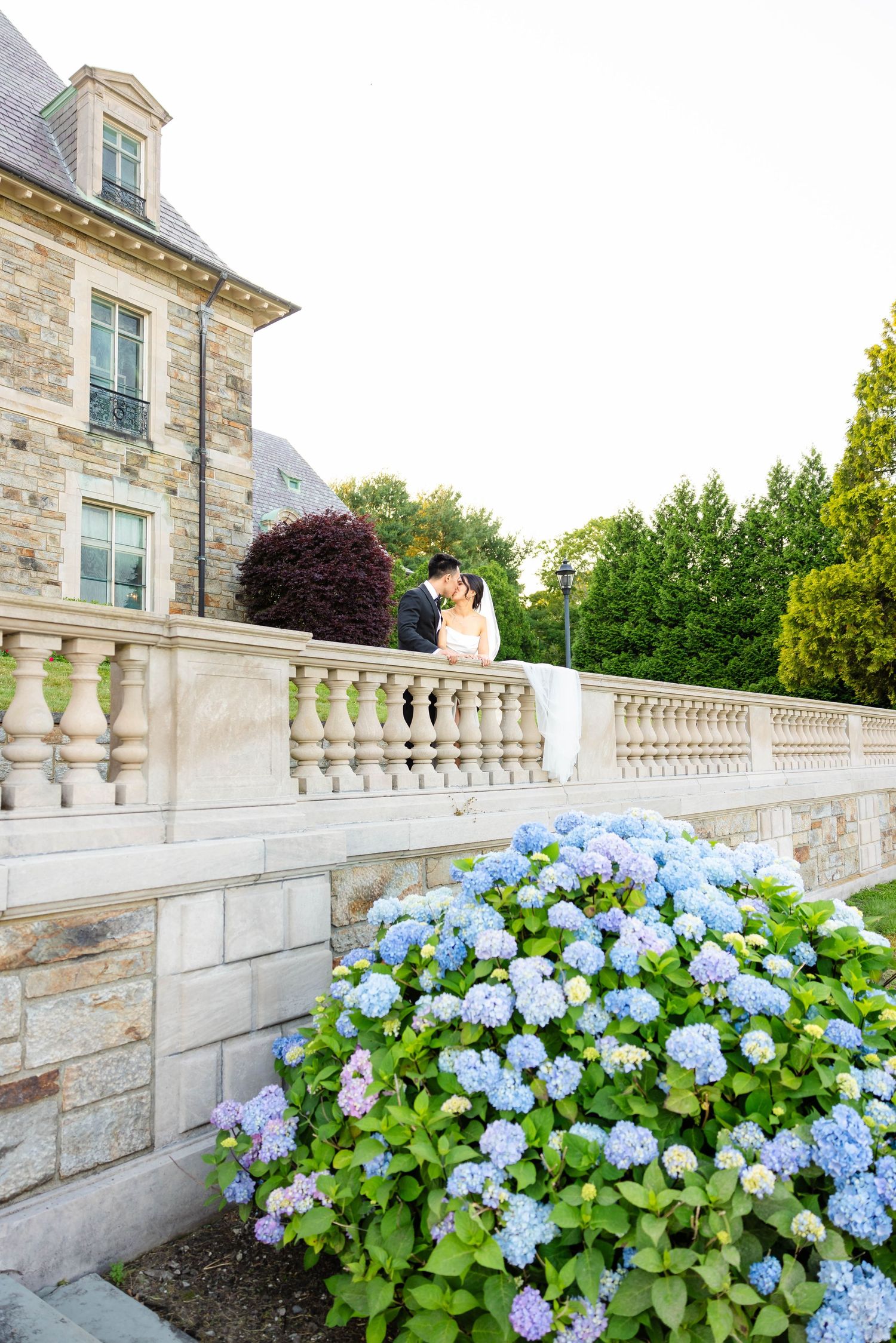 Bride and groom share a romantic kiss on a stone balustrade surrounded by blue hydrangeas at an elegant estate.
