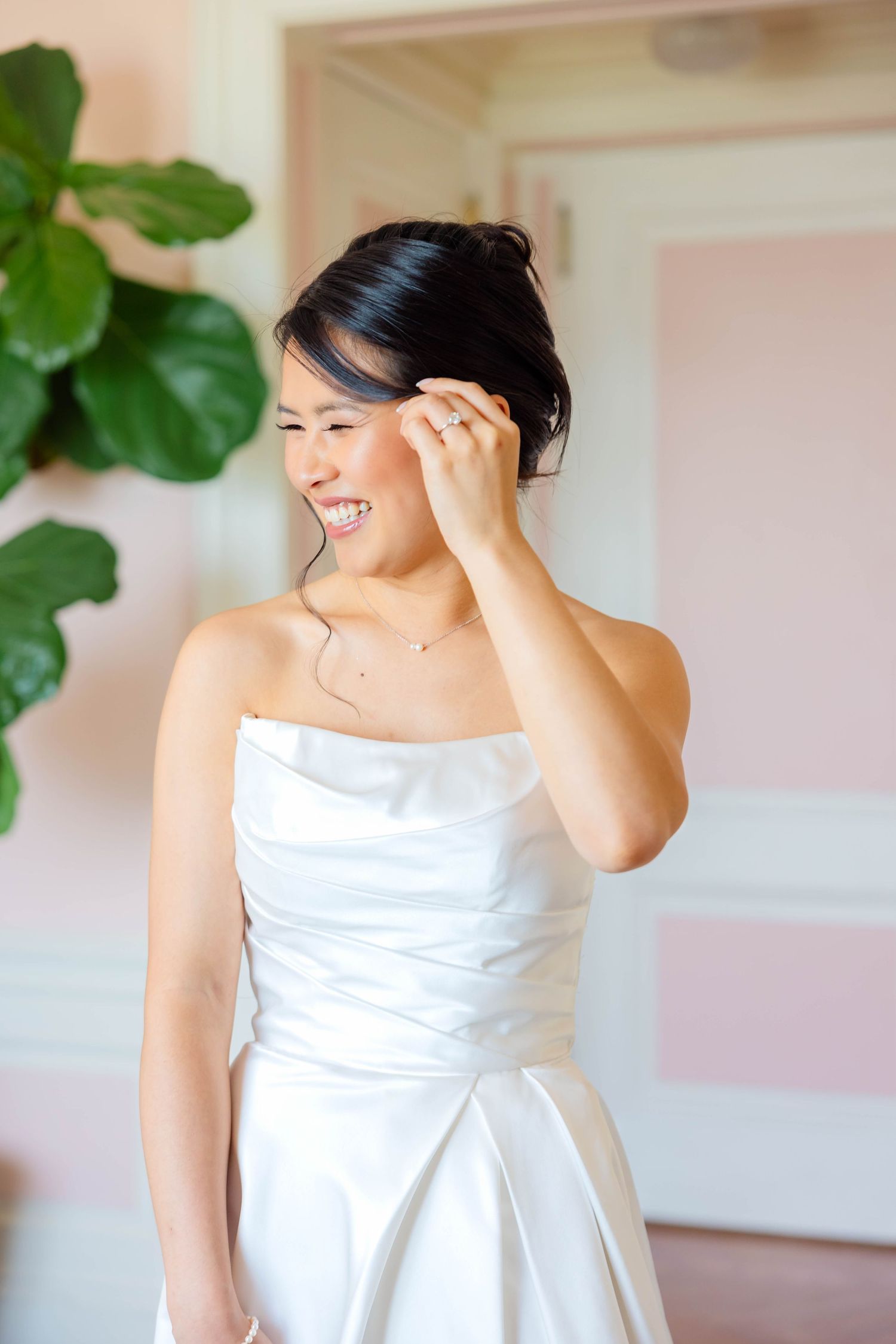 Smiling bride in strapless white gown touching her dark updo hair, standing in a softly lit pink room.