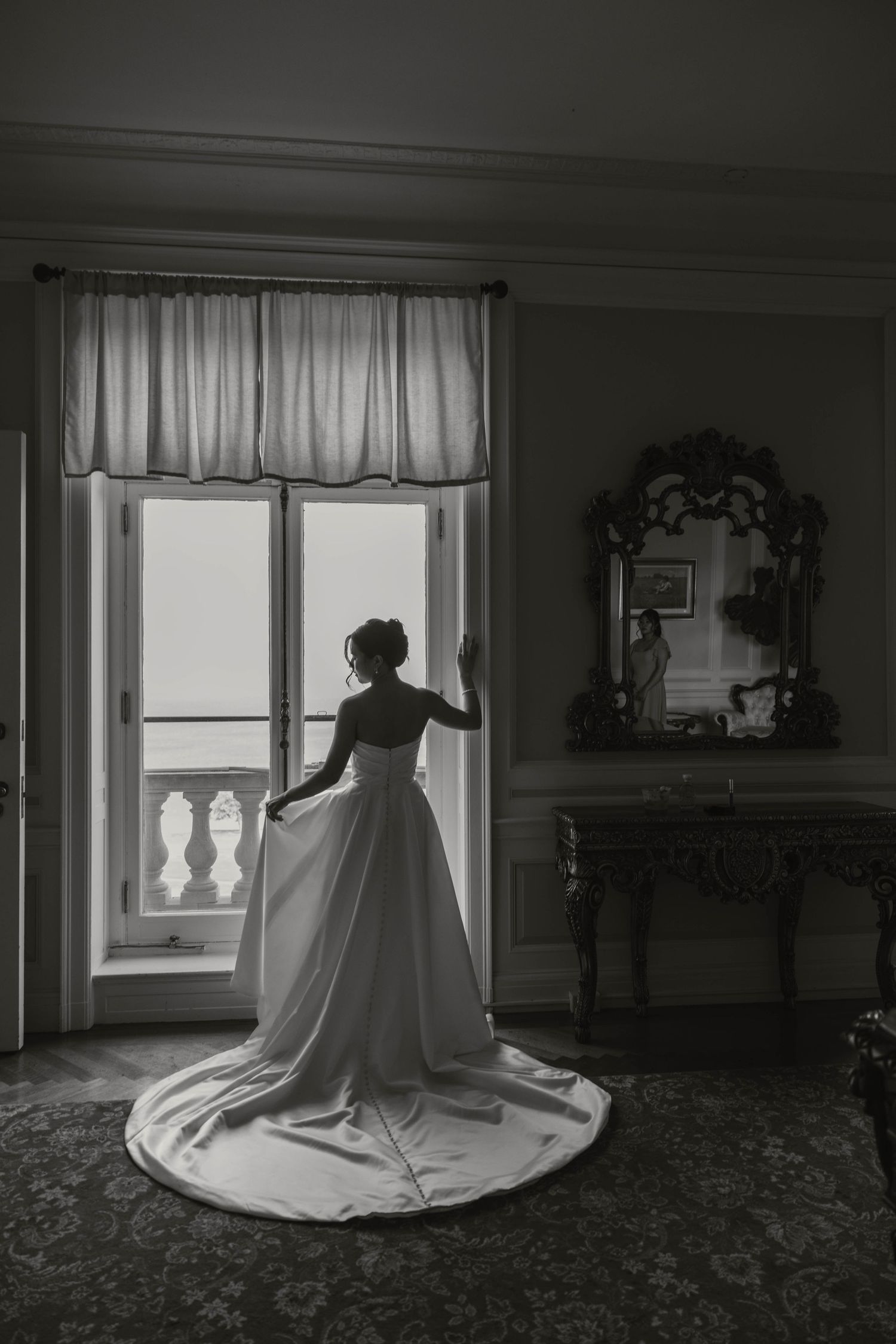 Bride in elegant white gown with long train stands at open French doors in black and white wedding photo.