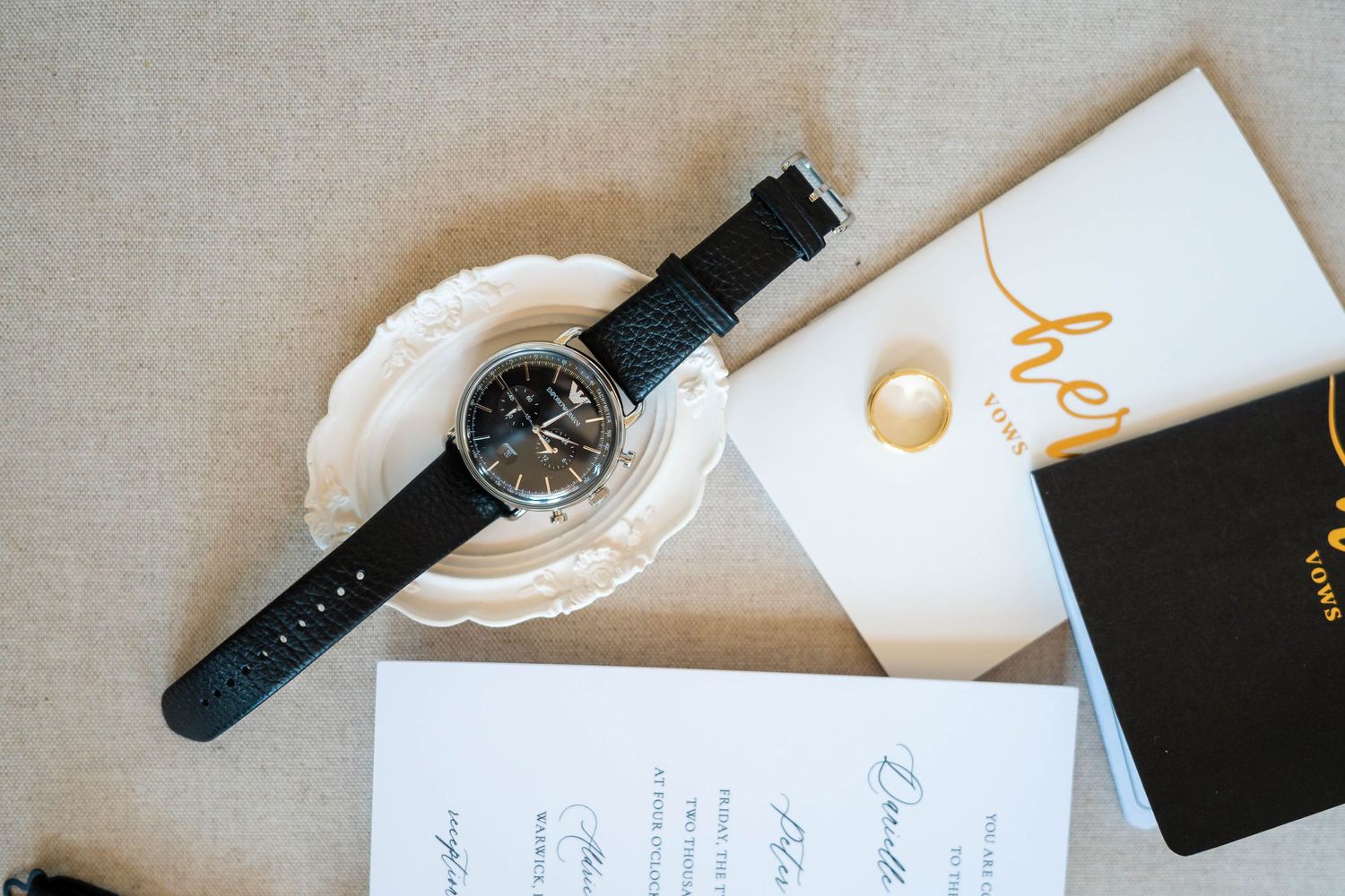Elegant wedding flat lay featuring a black leather watch, pearl earring, and script invitation on a neutral linen background.