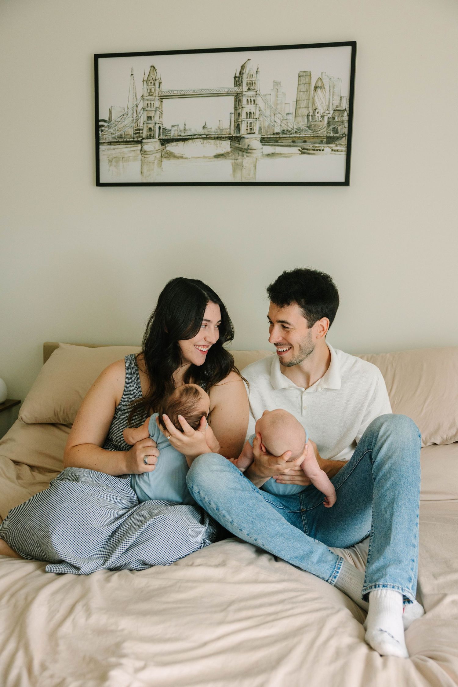 Happy young parents sit on a bed smiling at each other while holding their newborn baby together.