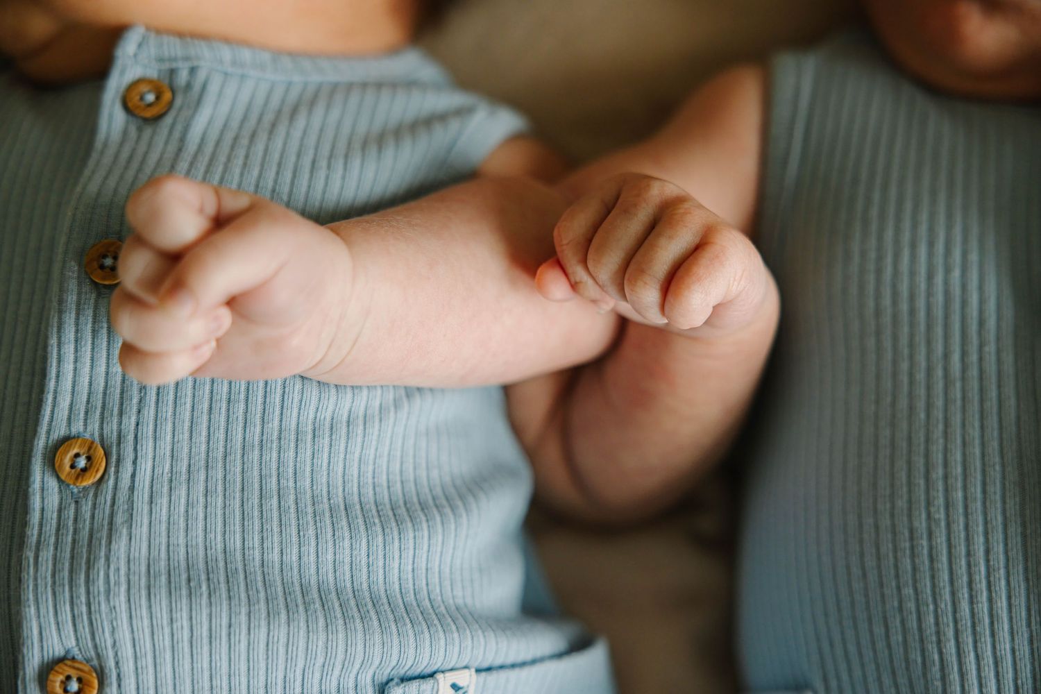 Close-up of a newborn baby's tiny hands resting on a light blue ribbed onesie with wooden buttons.