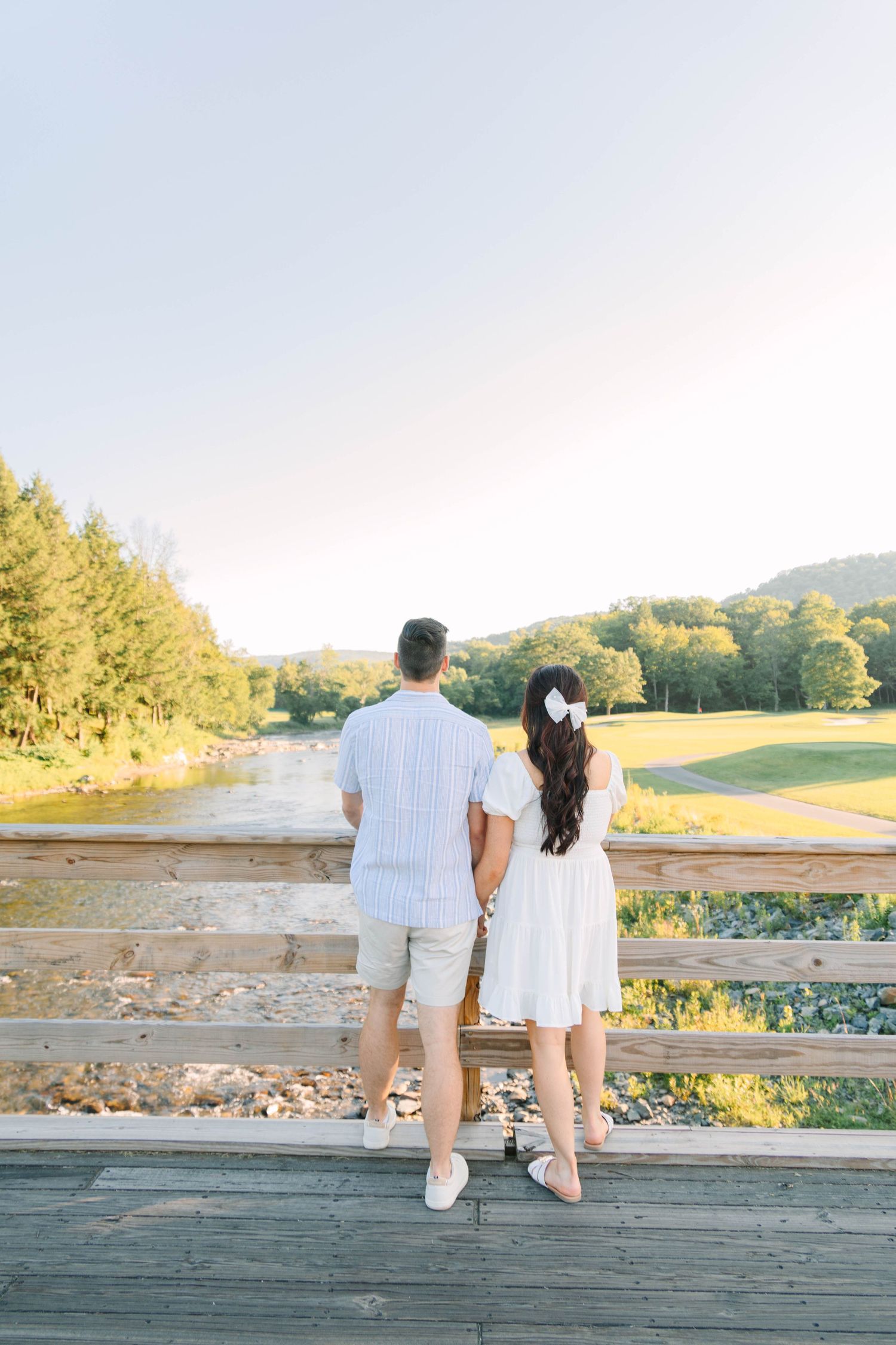 Couple holding hands on wooden bridge overlooking scenic river and green hills.