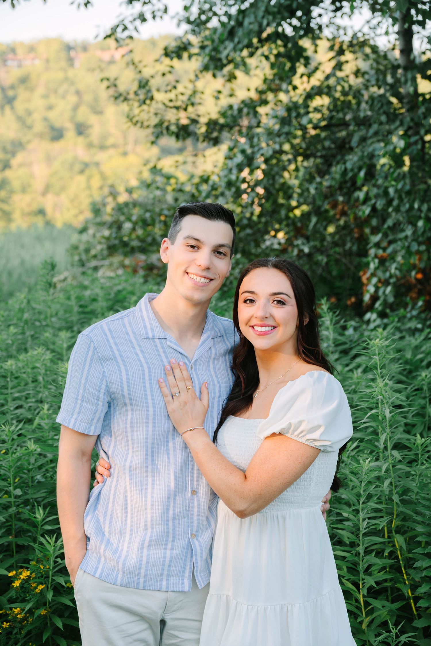 Happy engaged couple posing outdoors in lush greenery, woman showing engagement ring in white dress.