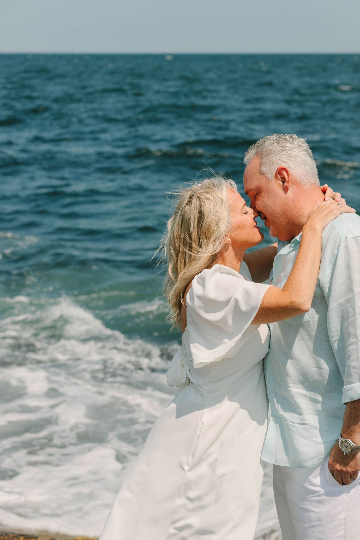Romantic senior couple kissing by the ocean waves, wearing white and light blue outfits.