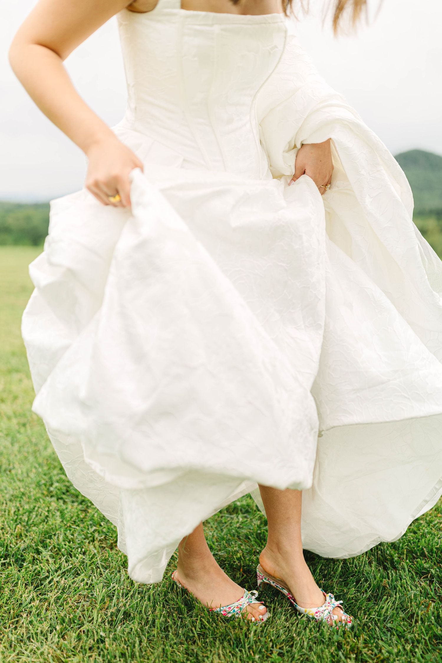 Close-up of bride lifting white wedding gown hem revealing silver embellished flat sandals on a green grass lawn.