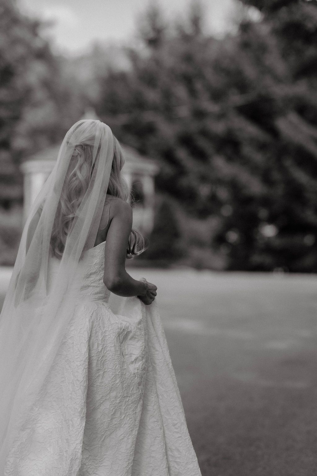 Black and white artistic photo of bride in lace gown and cathedral veil walking across a garden lawn.