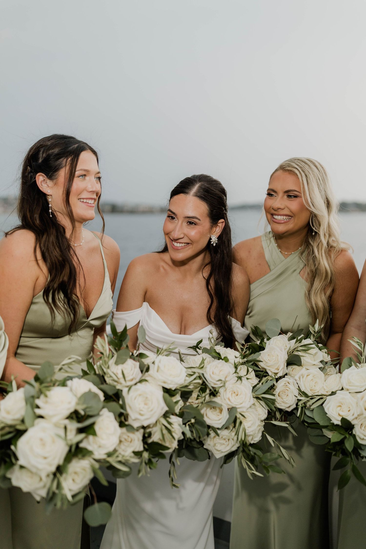 Bride and two bridesmaids in sage green dresses holding white rose bouquets near waterfront.