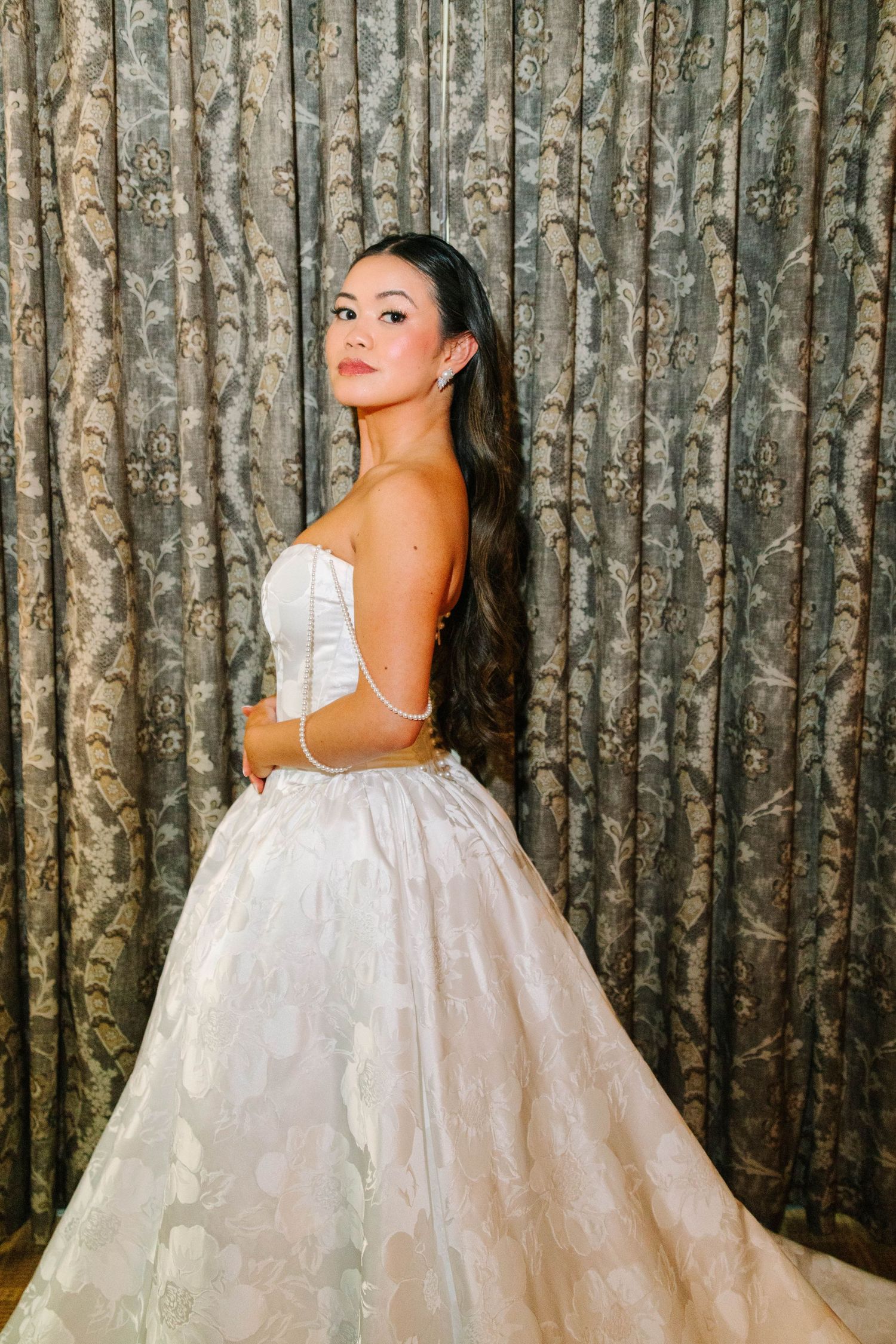 Bride in strapless white lace ballgown posing in front of elegant patterned curtains.