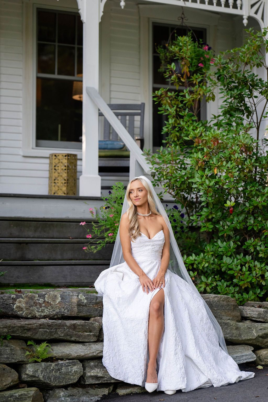 Bride in white lace gown with slit and veil sits on stone steps in front of a charming white Victorian house.