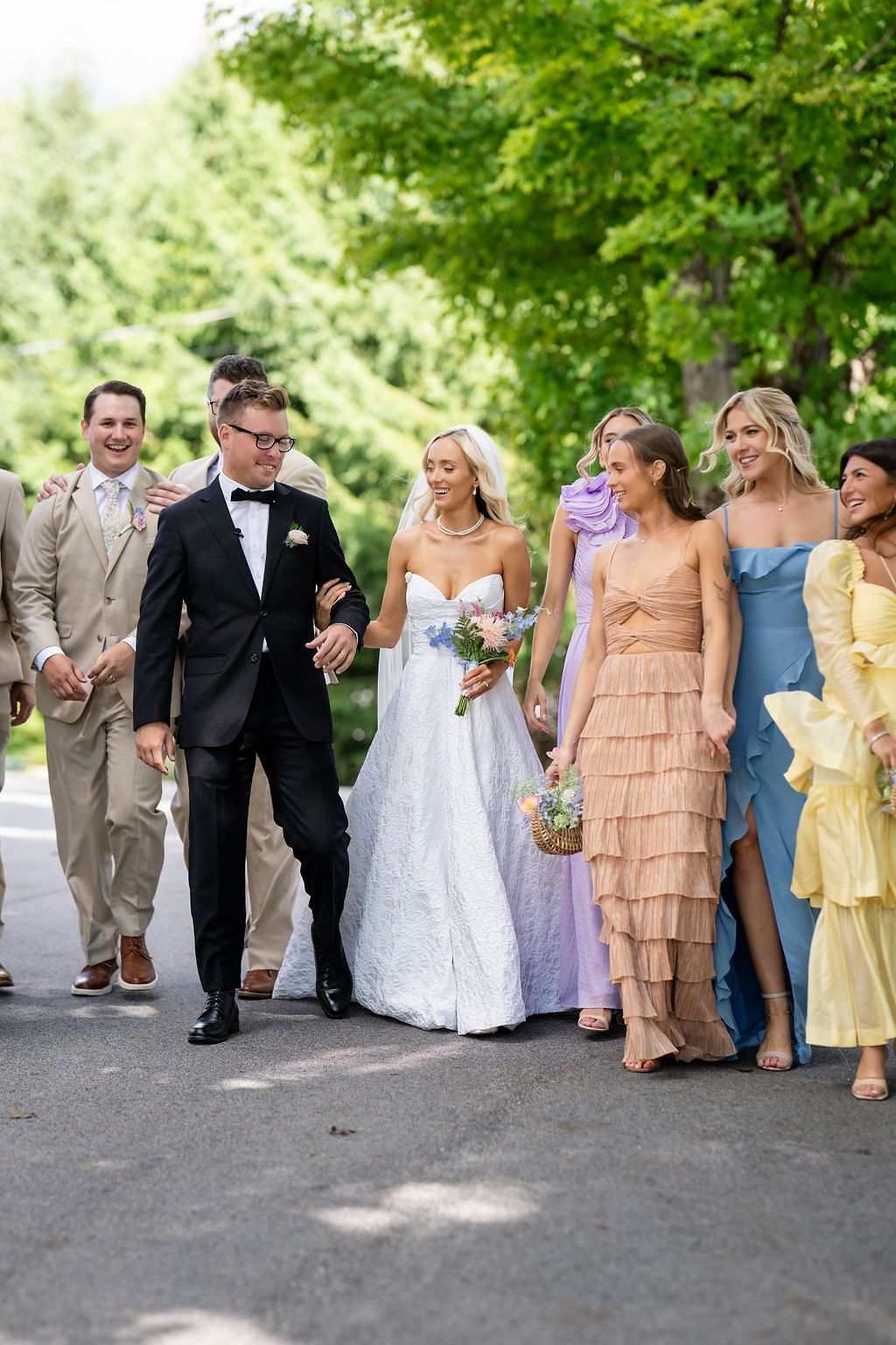 Wedding party walks outdoors, bride in white gown with colorful bridesmaids in pastel dresses alongside groomsmen.