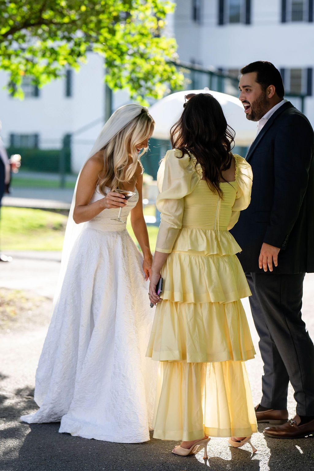 Bride in lace gown and veil shares joyful moment with bridesmaid in yellow dress and male guest.