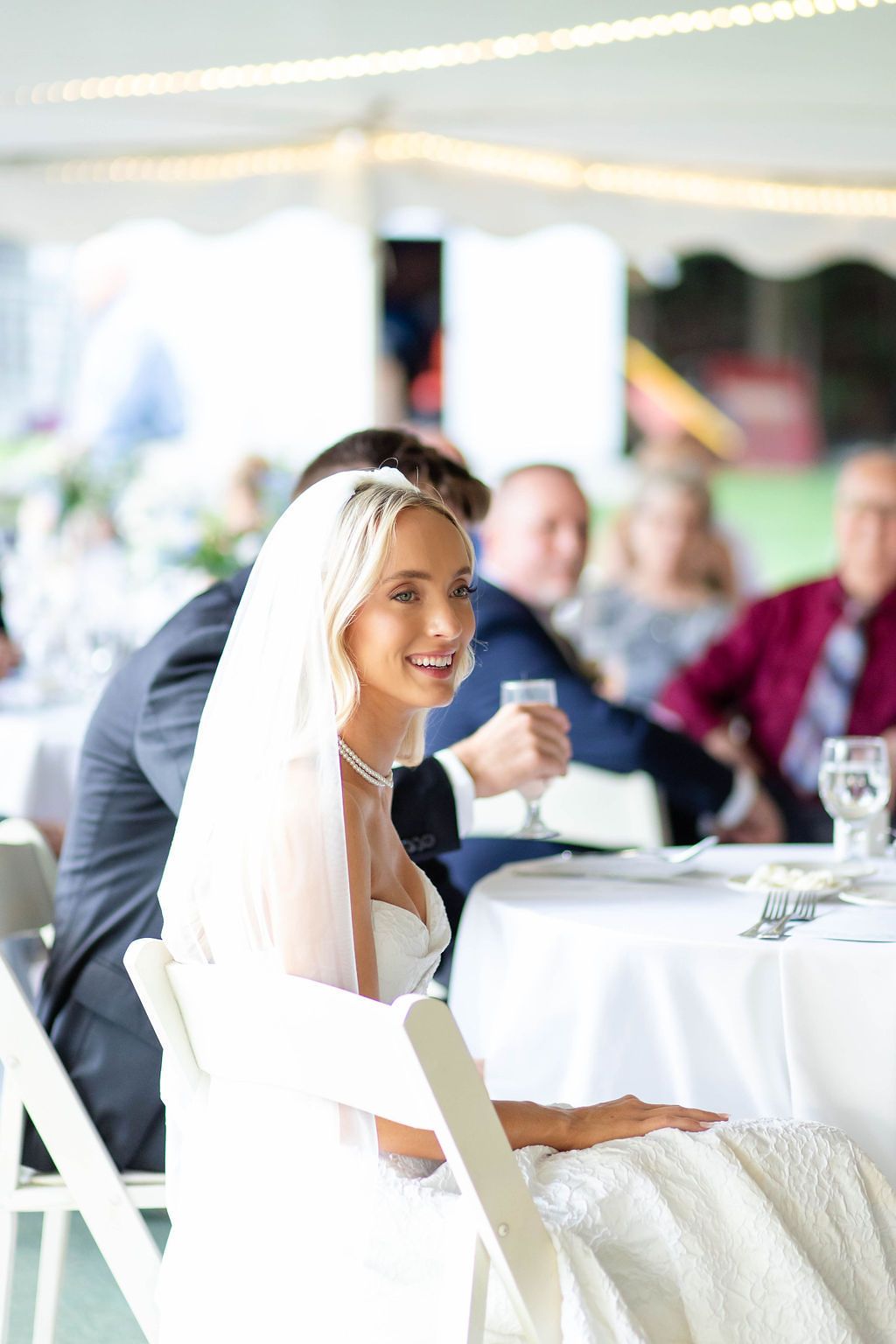 Smiling blonde bride in white gown and veil seated at wedding reception table with guests.