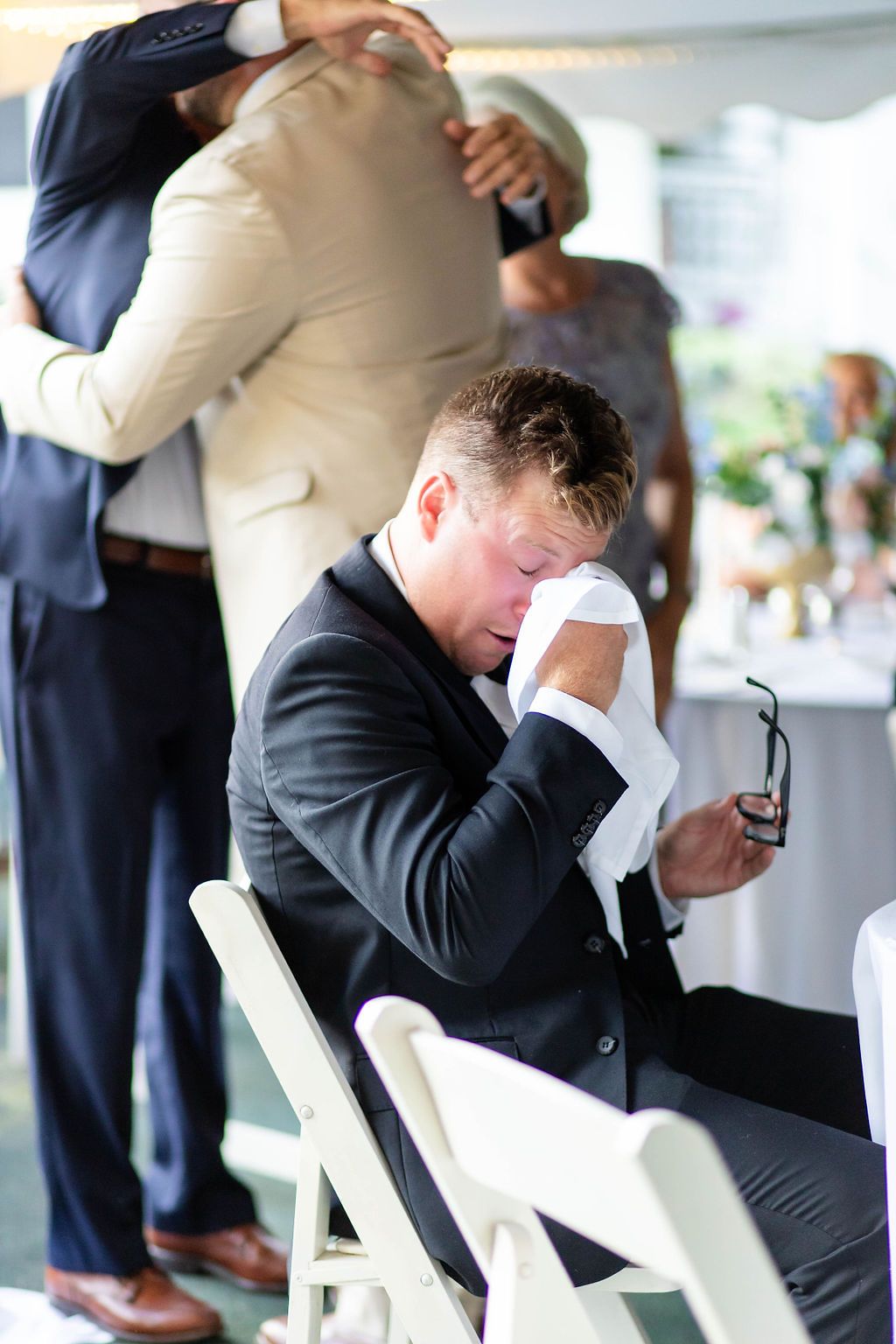 Emotional groom wipes tears with handkerchief while seated at wedding reception holding his glasses.