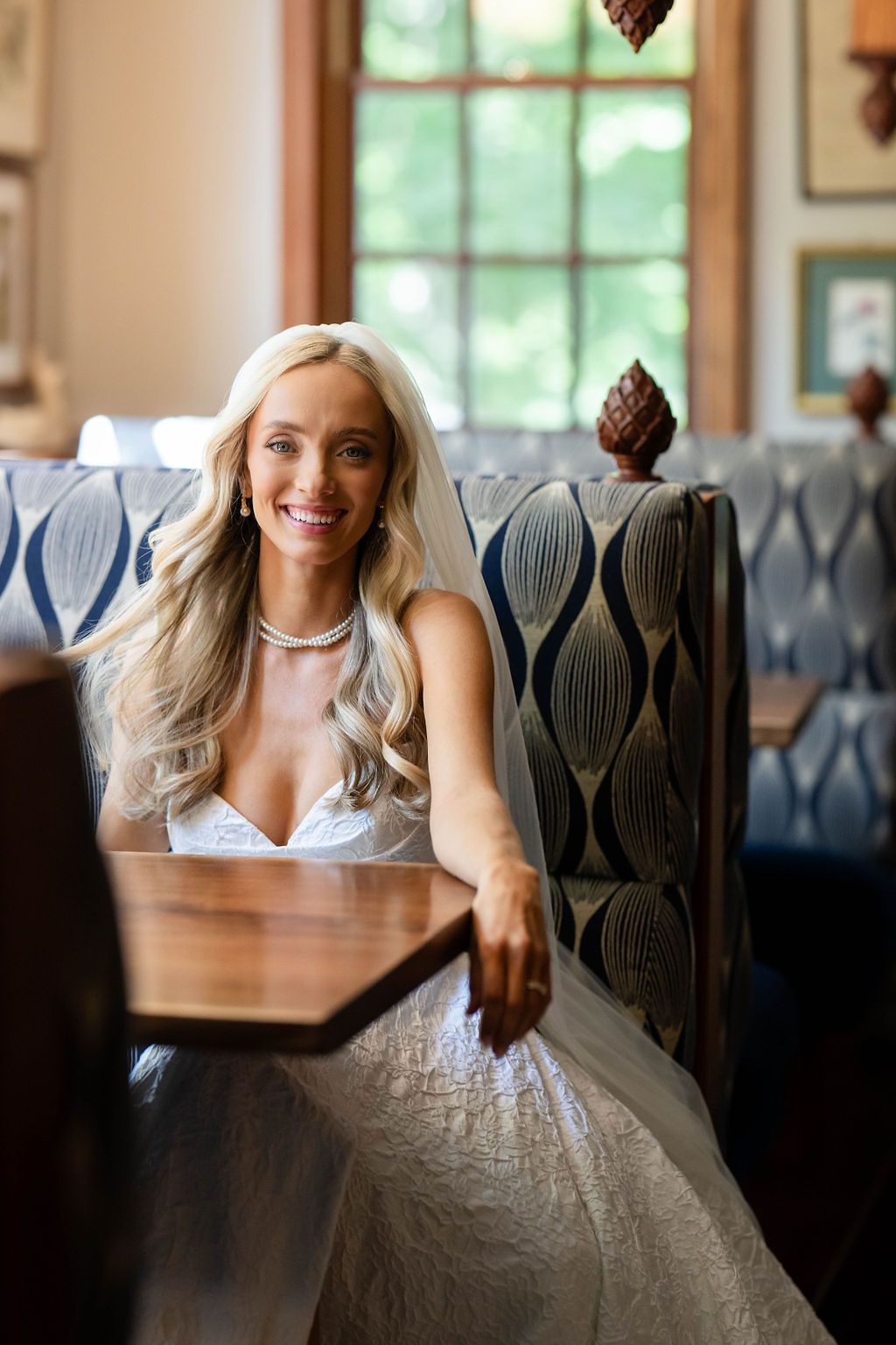 Smiling bride in a lace wedding gown and veil sits in an elegant booth inside a warmly lit restaurant.