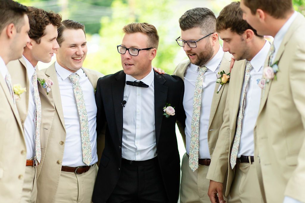 Groom in black tuxedo laughing and celebrating with groomsmen in tan suits at outdoor wedding.
