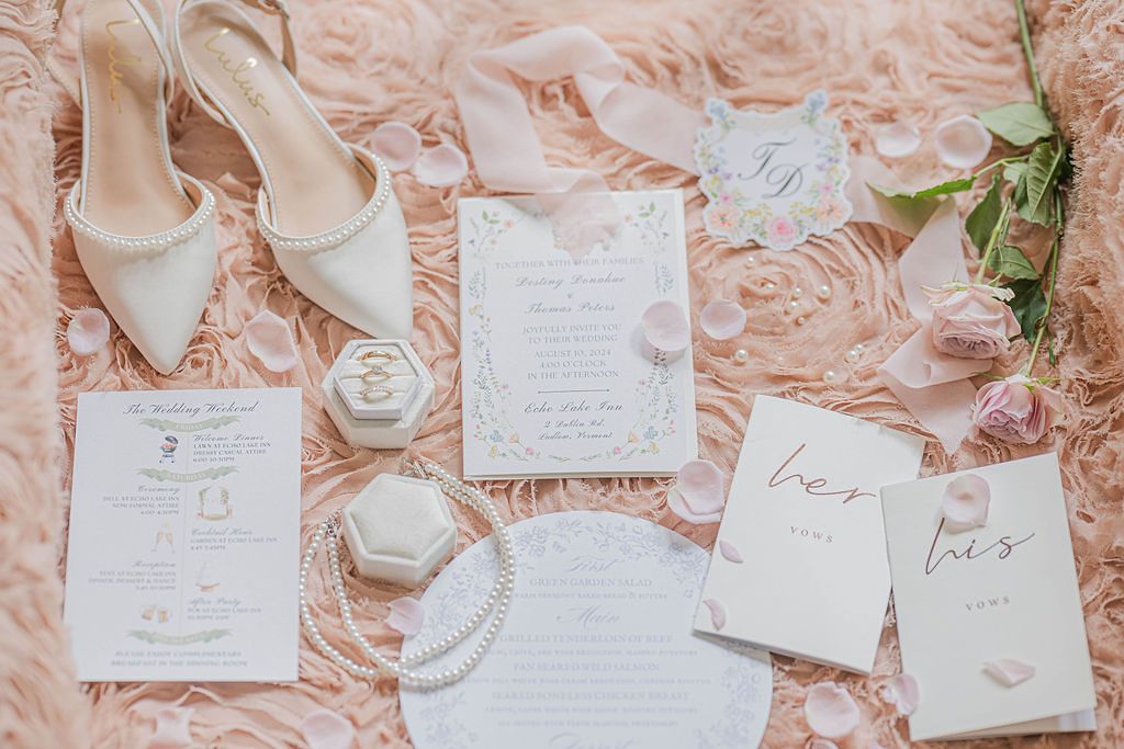 Elegant wedding flat lay with white heels, floral invitations, pearl jewelry, ring box, and pink roses on blush fur.