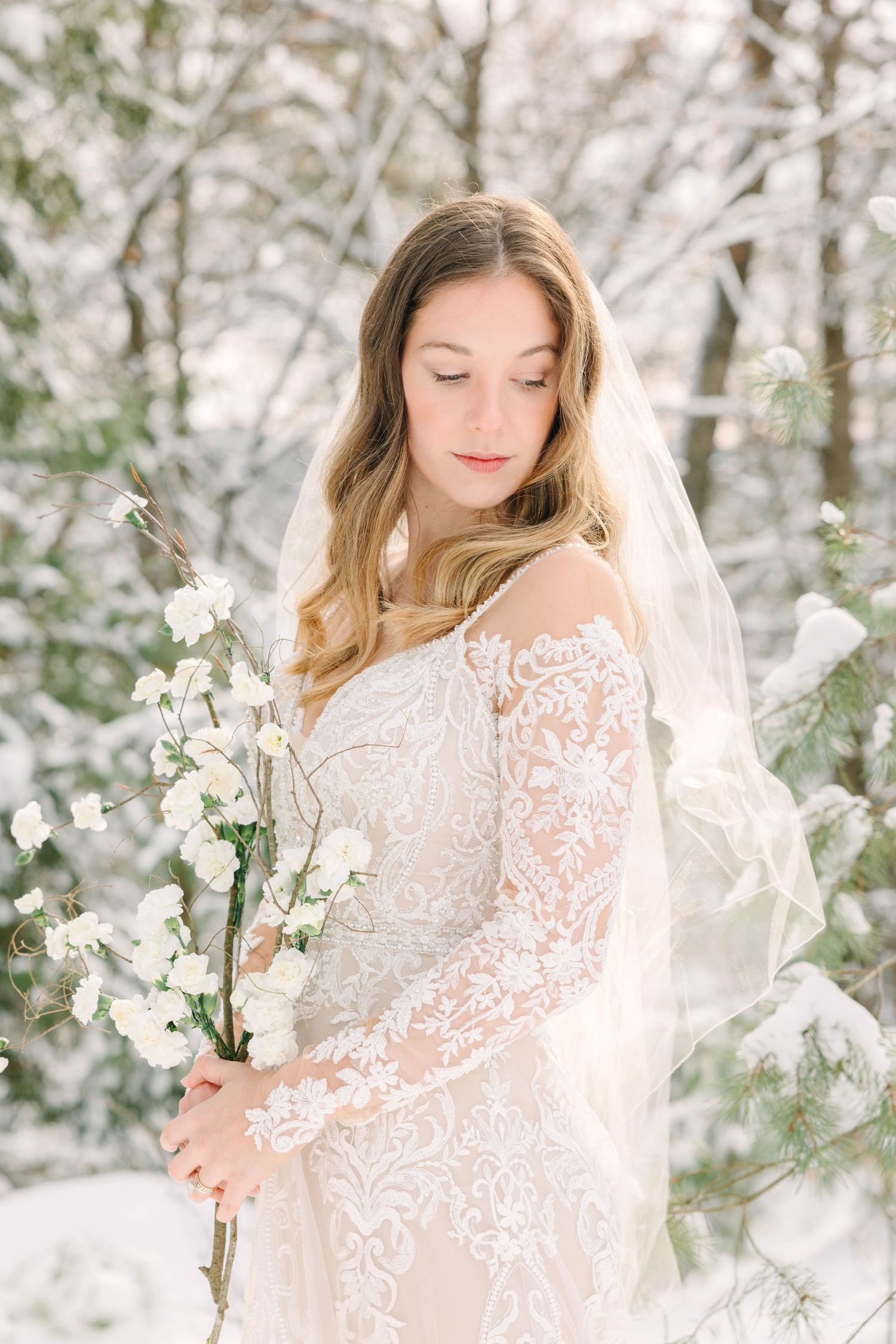 Bride in lace off-shoulder gown and veil holds white flowers in snowy winter forest setting.