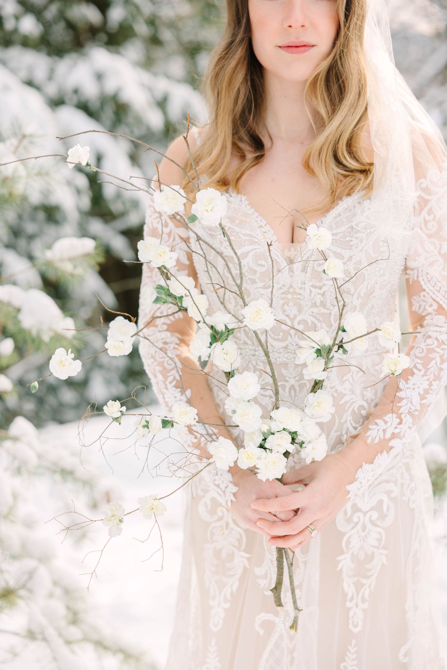 Bride in lace gown holding white floral branch bouquet in snowy winter forest.