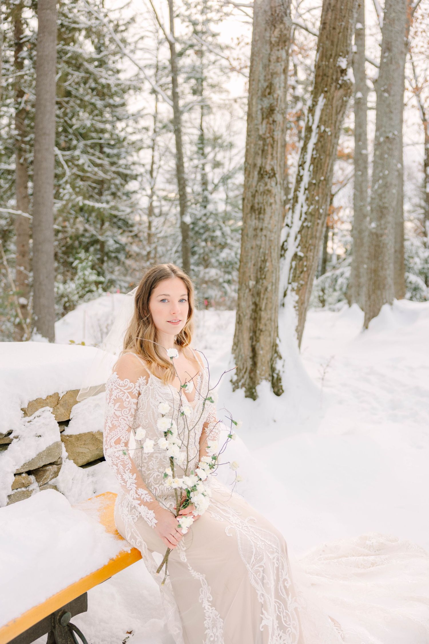 Smiling bride in lace wedding gown sits on snow-covered bench holding white flowers in a winter forest setting.