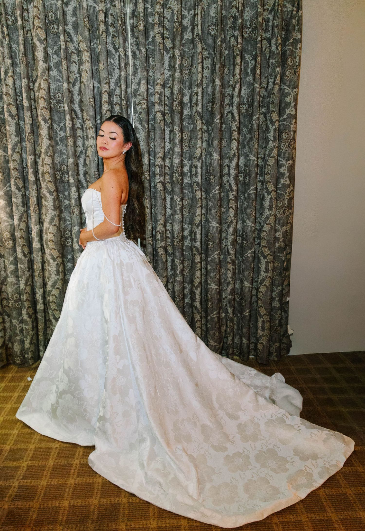 Bride in strapless white ballgown showing long flowing train before patterned curtain backdrop.