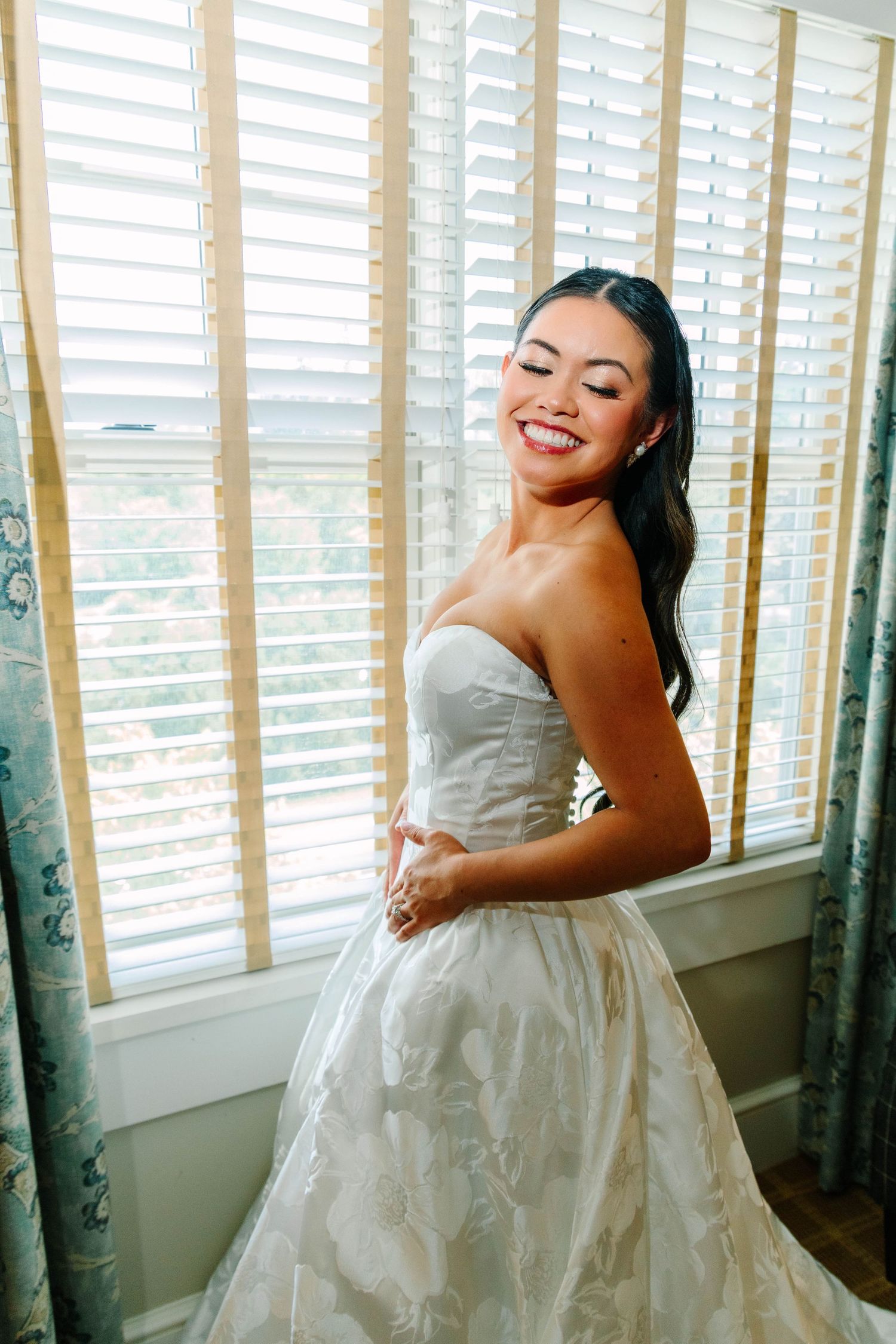 Radiant bride in strapless white ballgown smiling joyfully while posing in front of plantation shutters.