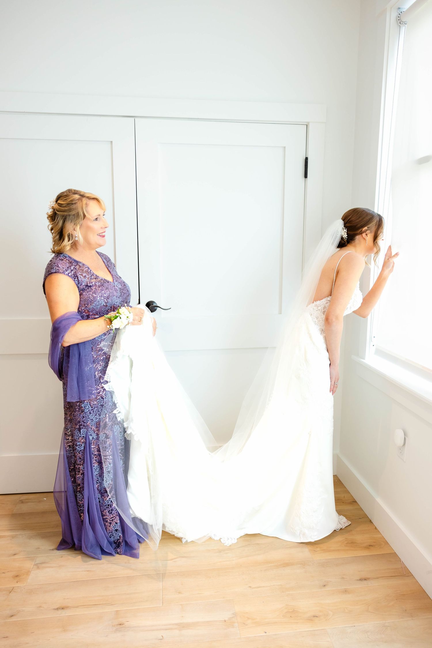Mother in purple gown holds bride's cathedral train as bride gazes out bright window indoors.