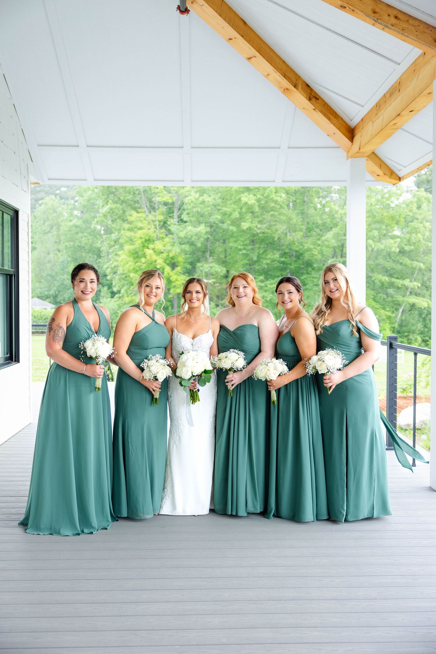 Bride and five bridesmaids in sage green gowns hold white rose bouquets on covered porch.