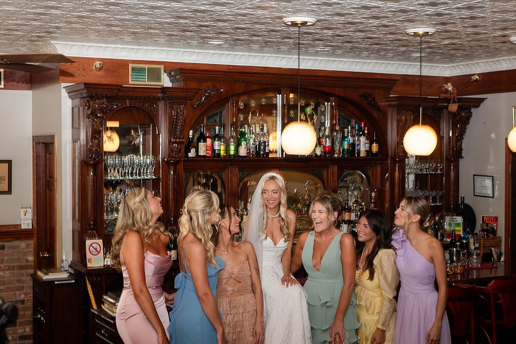 Bride in white gown laughs with bridesmaids in colorful dresses gathered around ornate wooden bar.