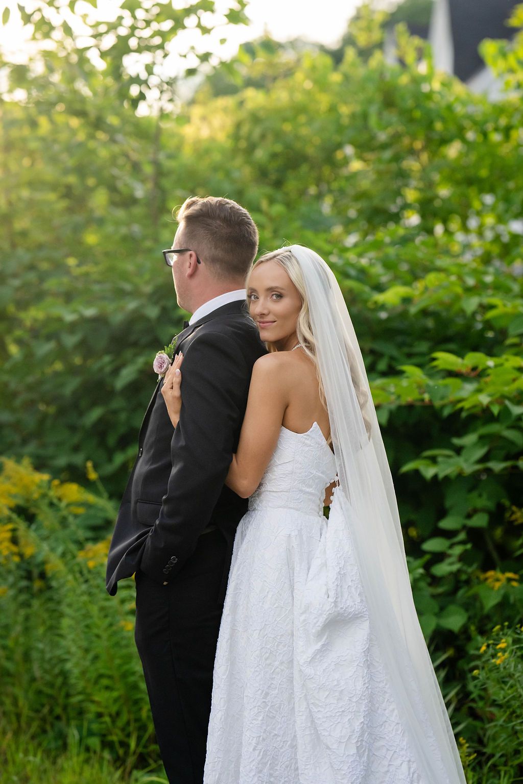 Bride in white lace gown and veil embraces groom in black suit outdoors among lush green foliage.