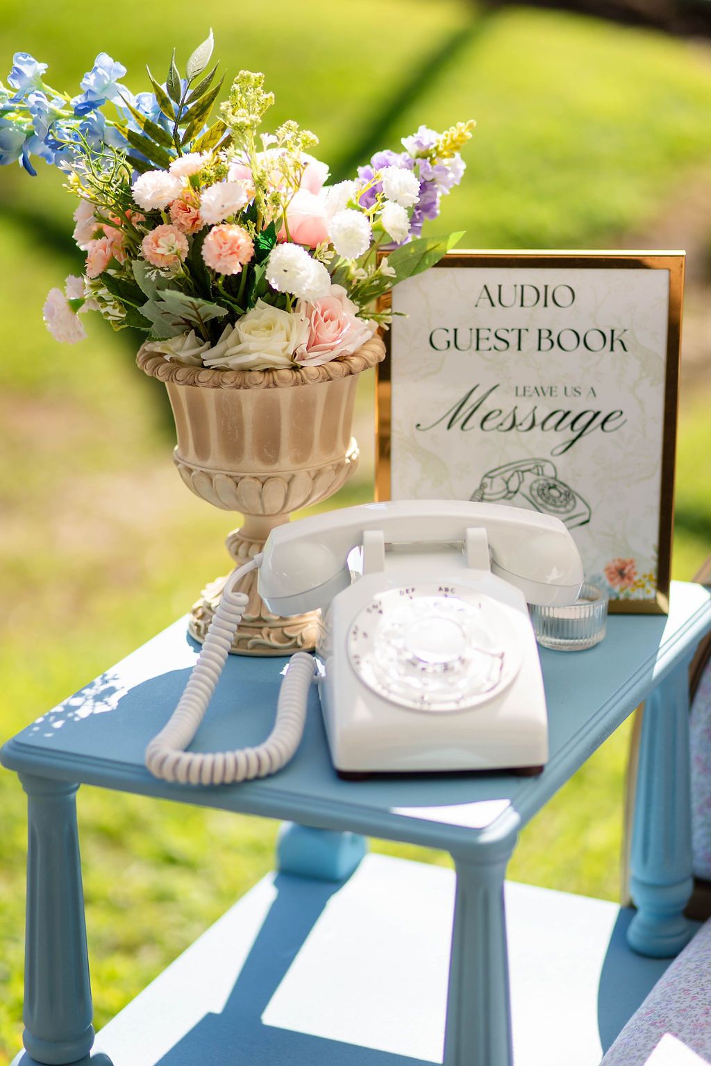 Vintage white rotary phone audio guestbook on blue table with floral arrangement at outdoor wedding.