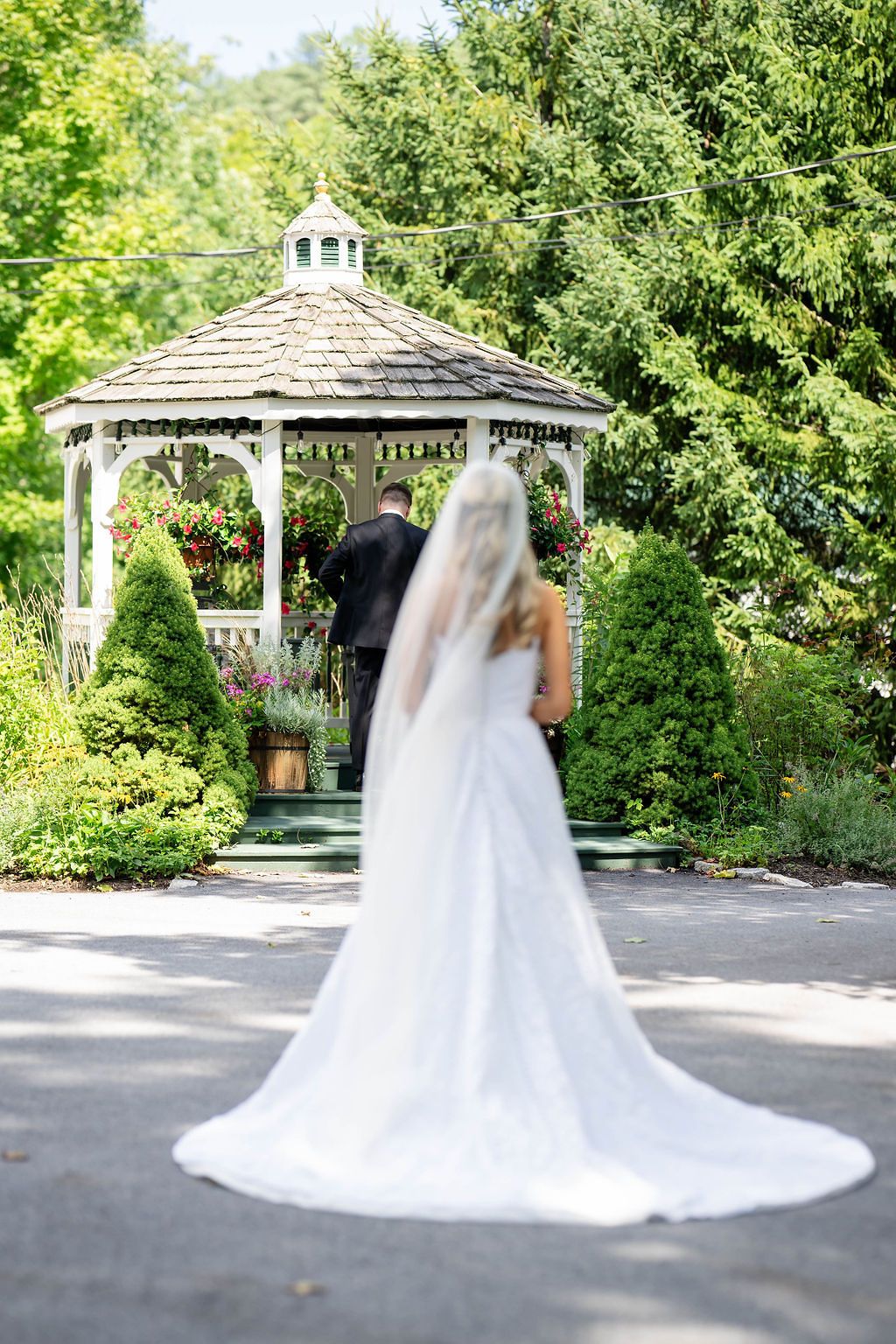 Bride in white gown with long veil walks toward groom at white gazebo surrounded by lush greenery.