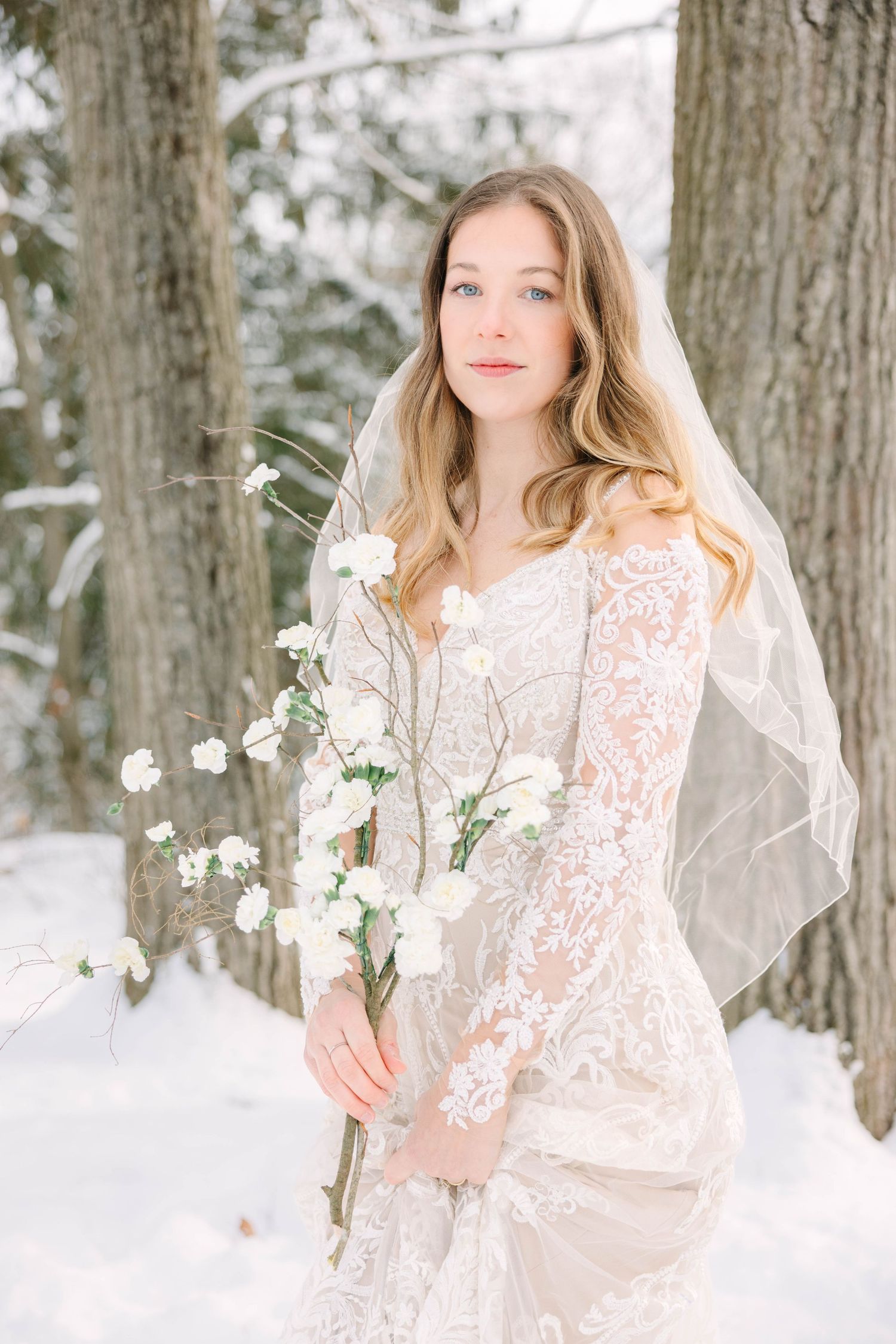 Blue-eyed bride in lace wedding gown and veil poses with white bouquet among snow-dusted trees.