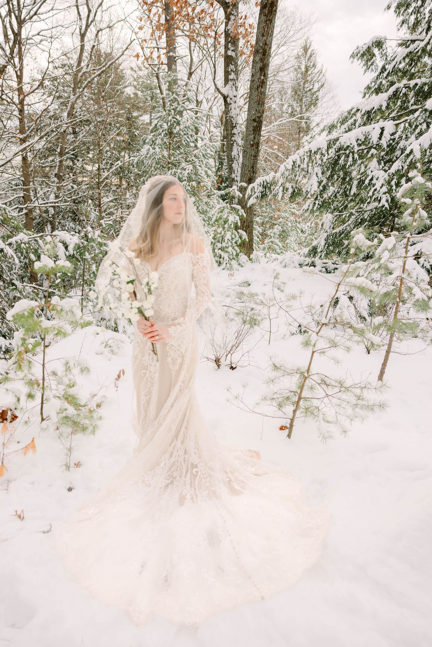 Full-length view of bride in lace wedding dress and veil standing in deep snow-covered winter forest.