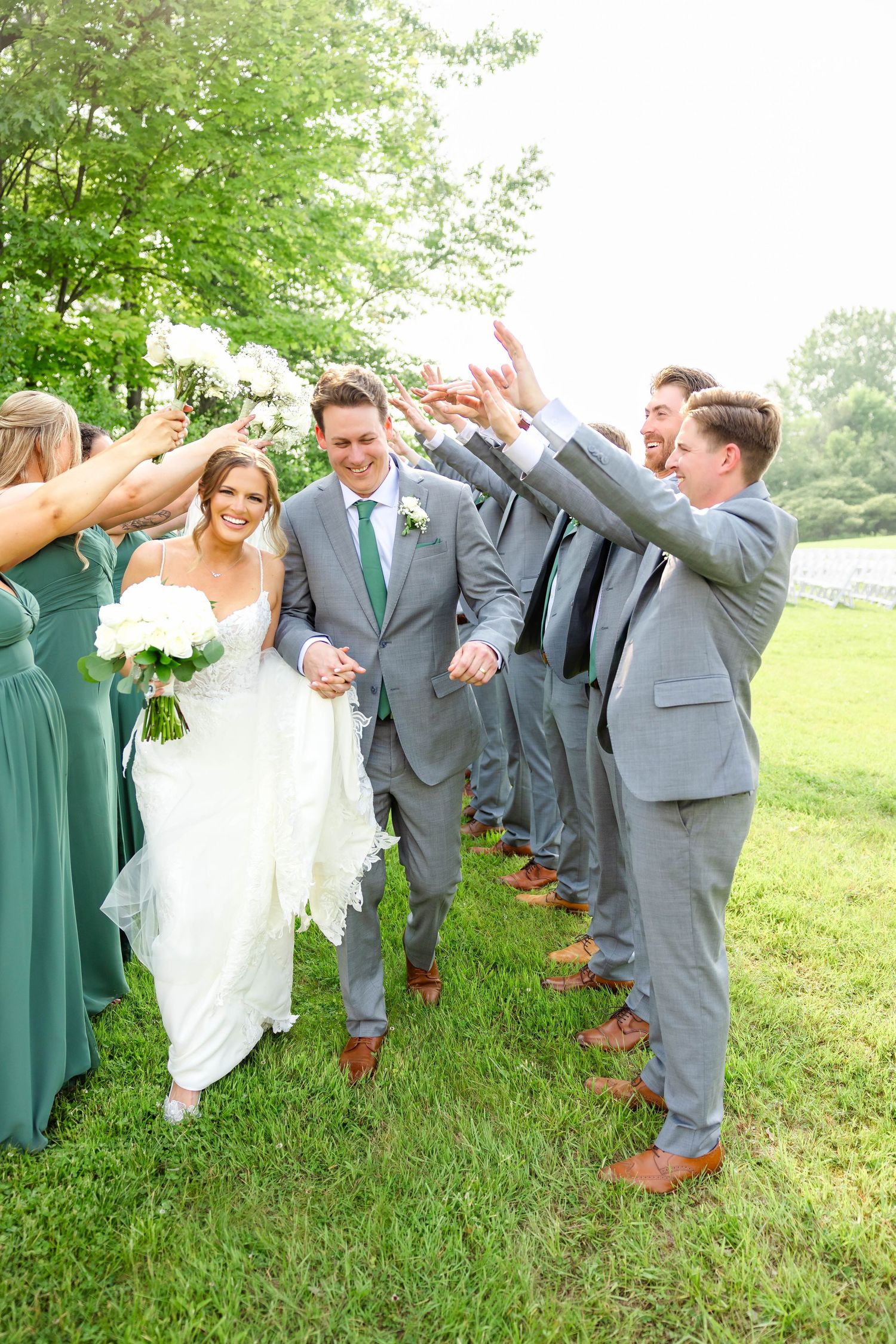 Bride and groom walking through arch of raised hands formed by wedding party on green lawn.