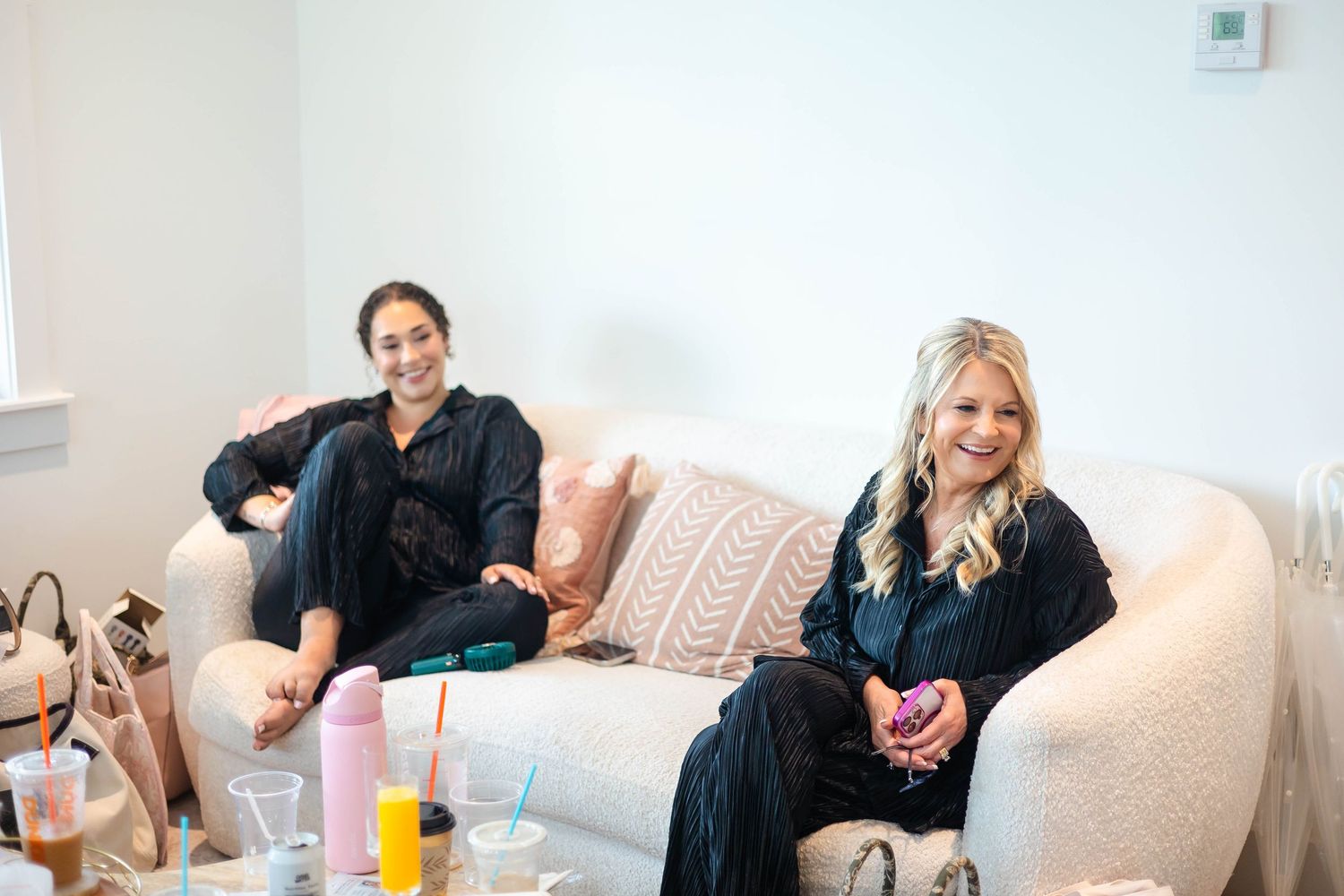 Two bridesmaids in black robes relaxing on a white couch surrounded by drinks and bags before the wedding.