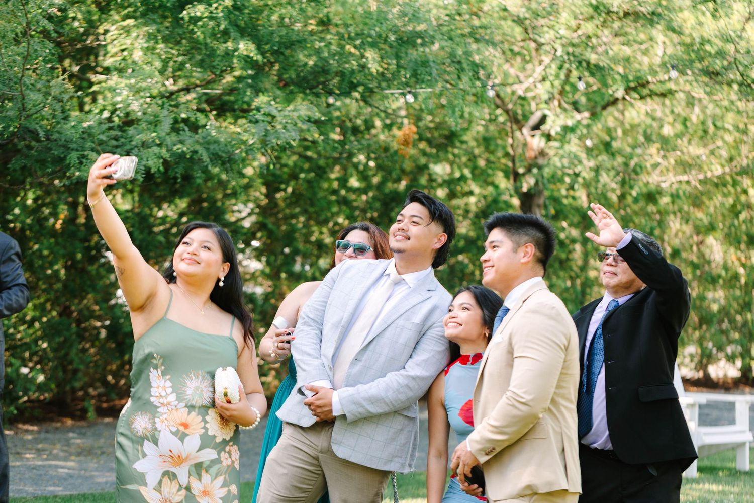 Wedding guests taking a group selfie outdoors surrounded by lush green trees at a wedding celebration.