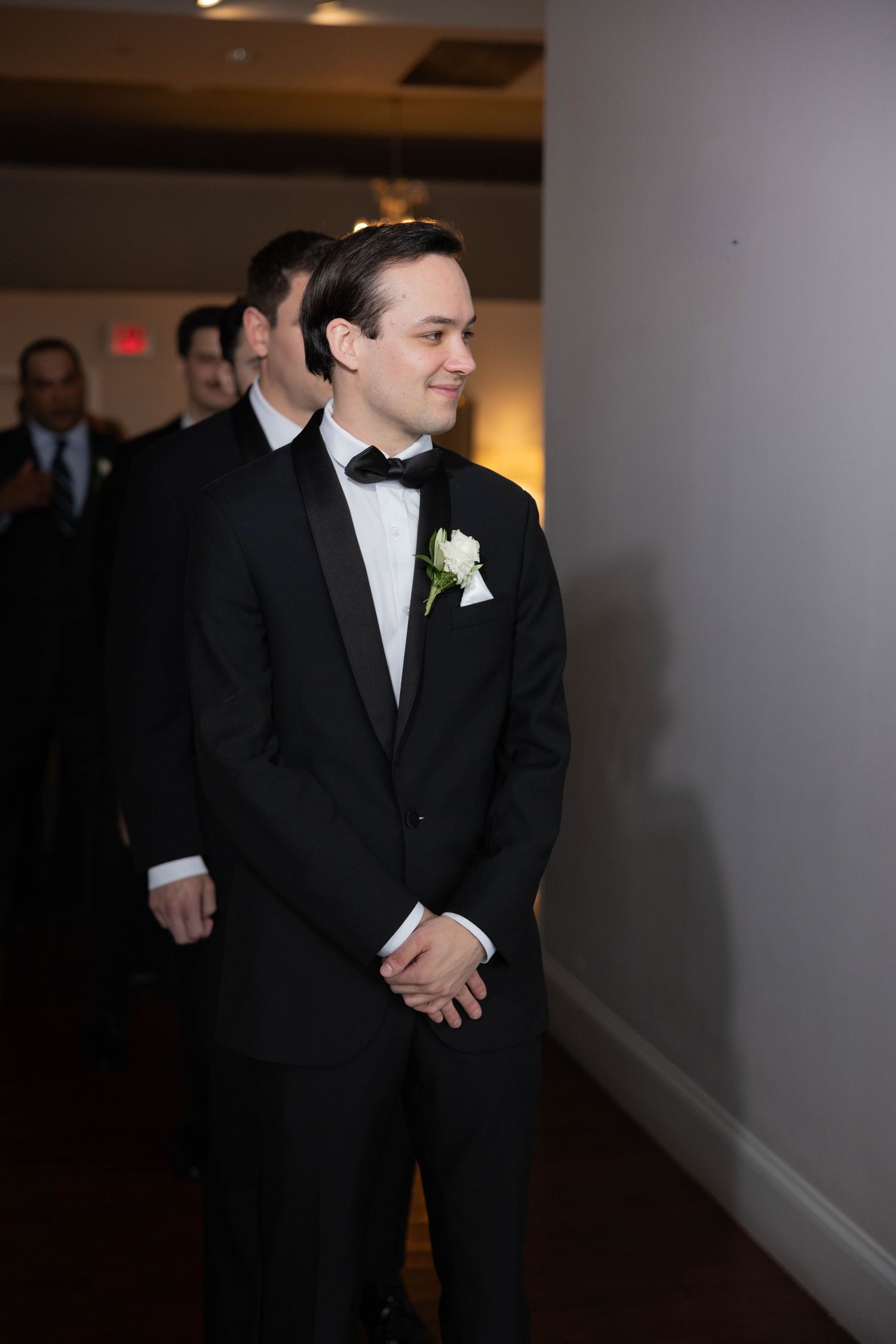 Groom in black tuxedo smiling while waiting at wedding ceremony entrance.
