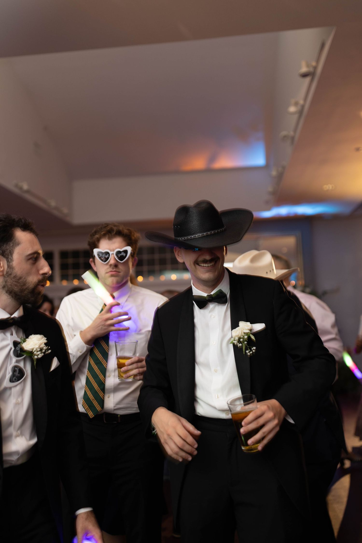 Groomsmen in tuxedos celebrate at a wedding reception, one wearing a cowboy hat and another in heart-shaped sunglasses.