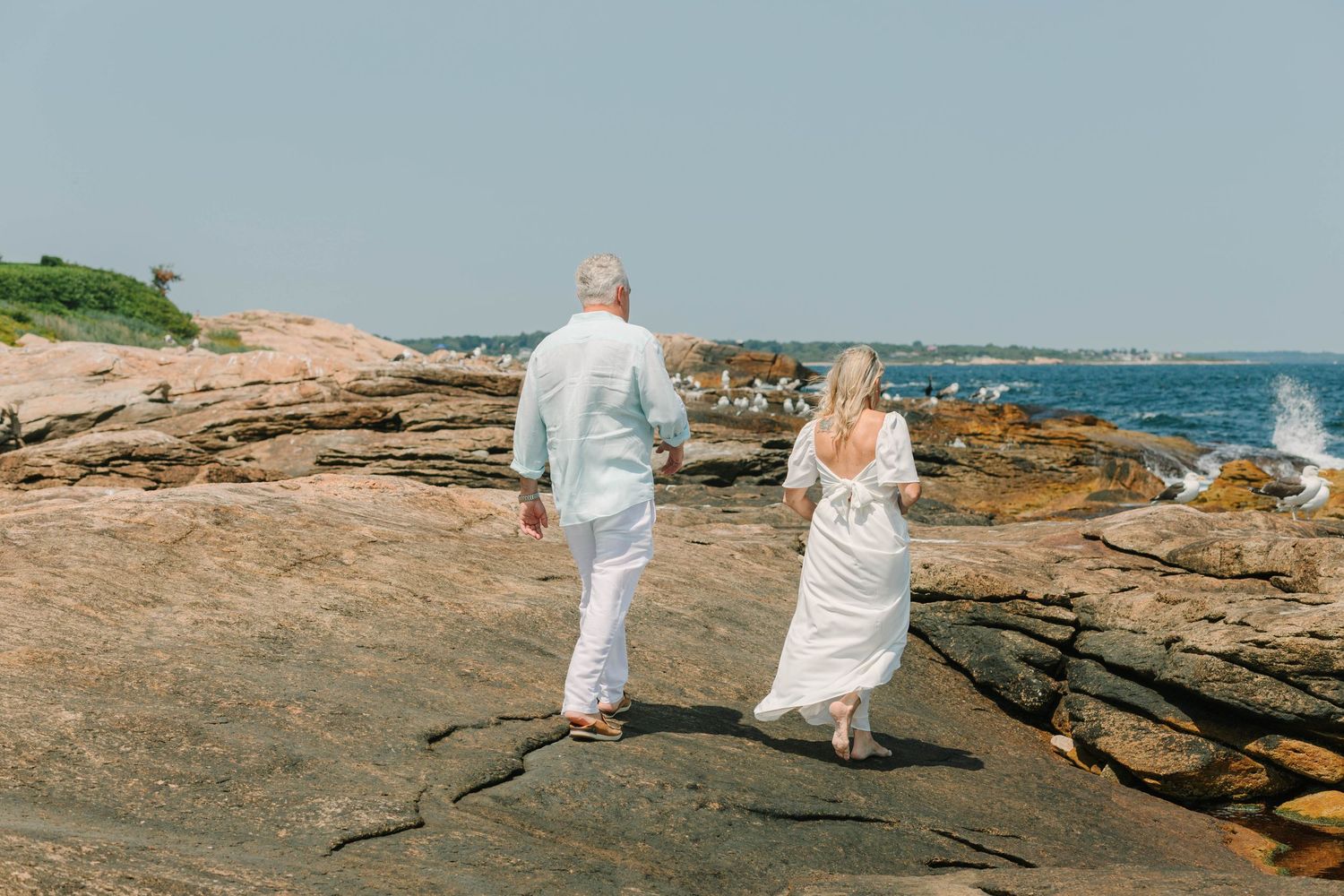 Couple in white attire walking hand in hand on rocky coastal shoreline with ocean views.