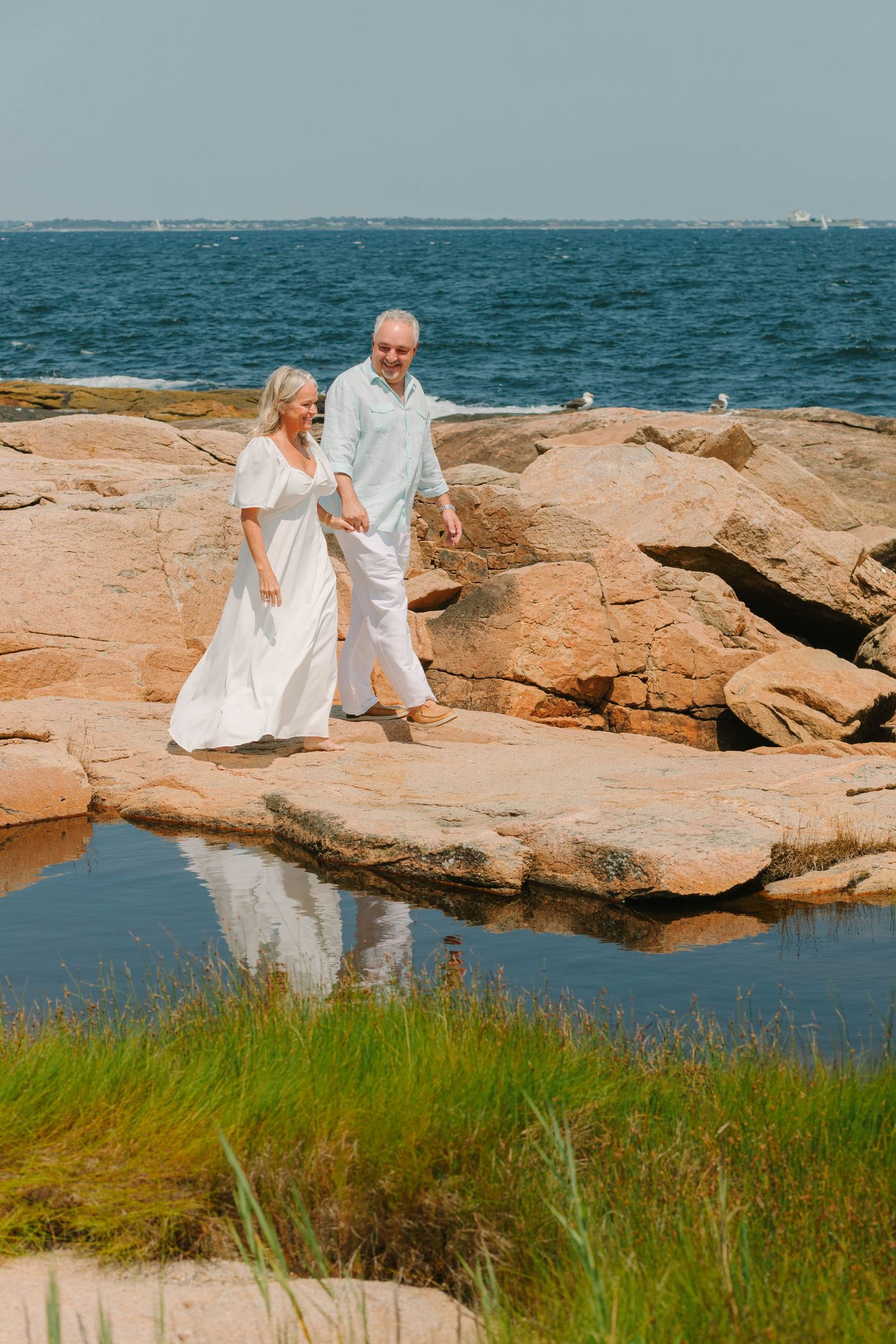 Elderly couple in white outfits walking hand-in-hand on rocky coastal shore with blue ocean in background.