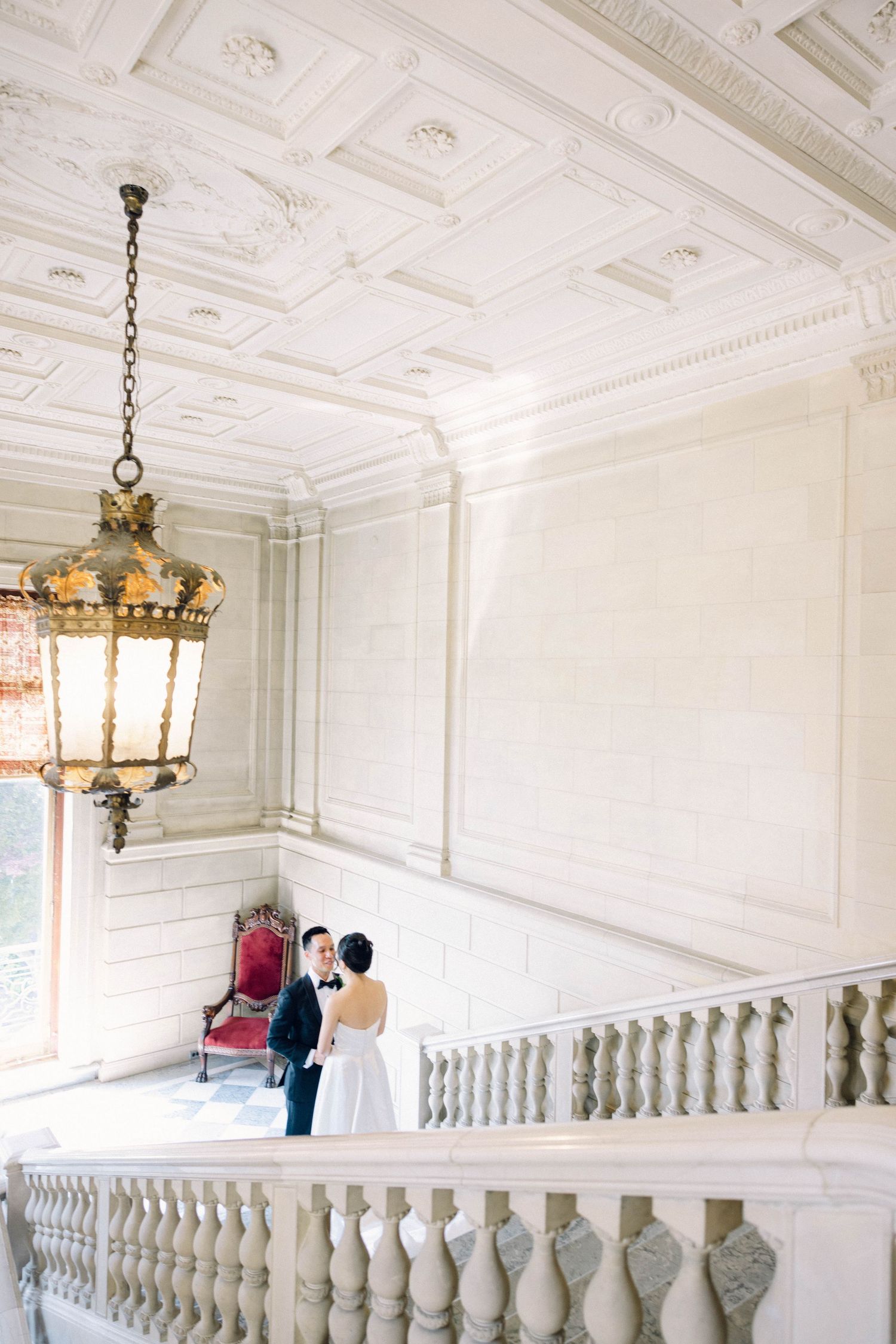 Elegant bride and groom portrait on grand marble staircase beneath ornate coffered ceiling with vintage chandelier.