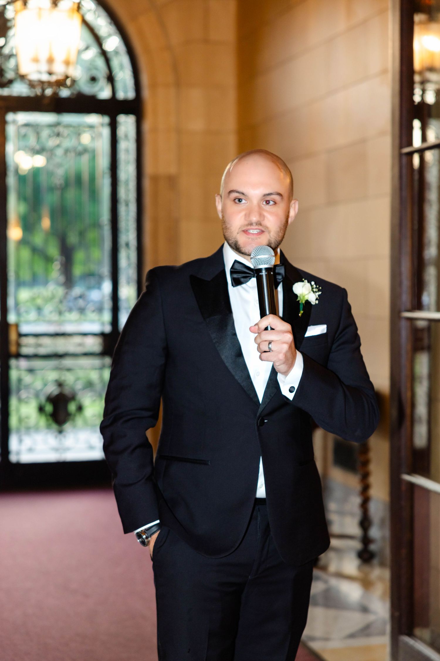 Bald groom in black tuxedo speaks into microphone during elegant wedding reception speech.