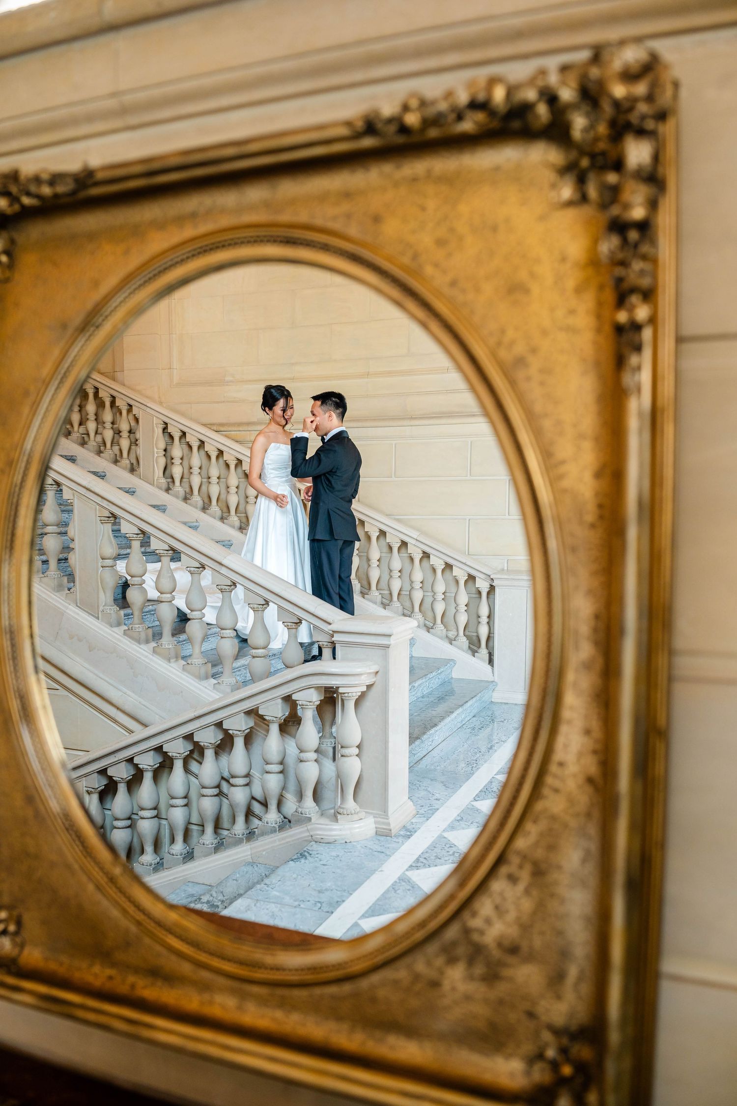 Bride and groom reflected in ornate gold oval mirror frame on grand marble staircase.